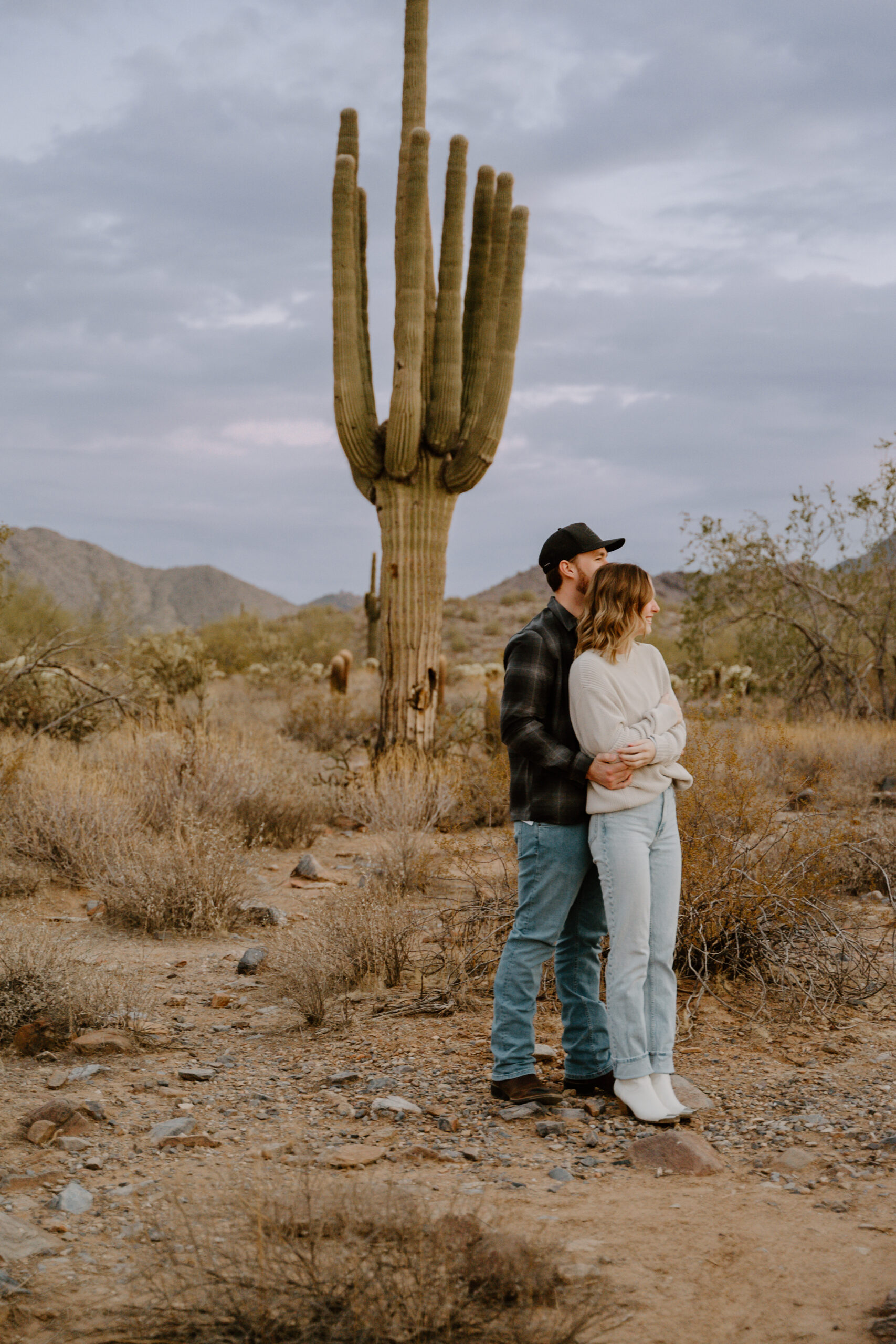 A couple stands in the Arizona desert, embracing in front of a towering saguaro cactus. The golden hour light highlights the natural beauty of Arizona engagement session locations.