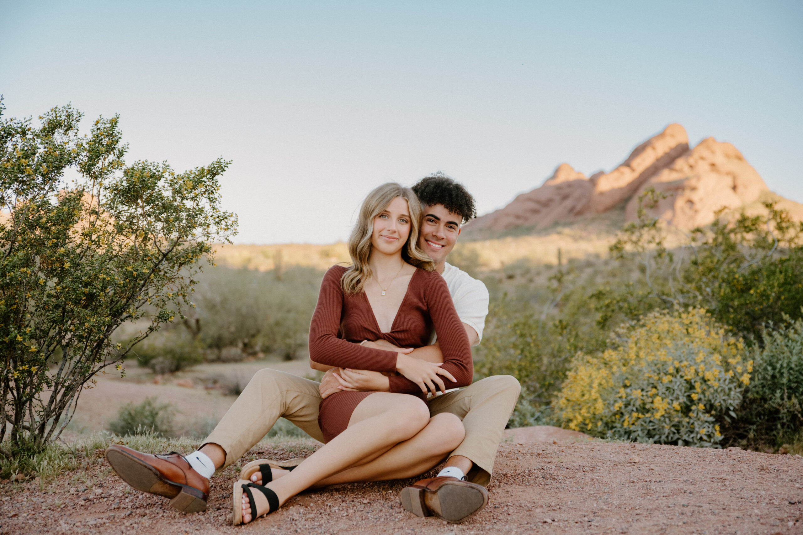 Couple sitting together in front of Papago Park's red rock formations and palm trees, showcasing one of the most diverse and scenic Arizona engagement session locations.