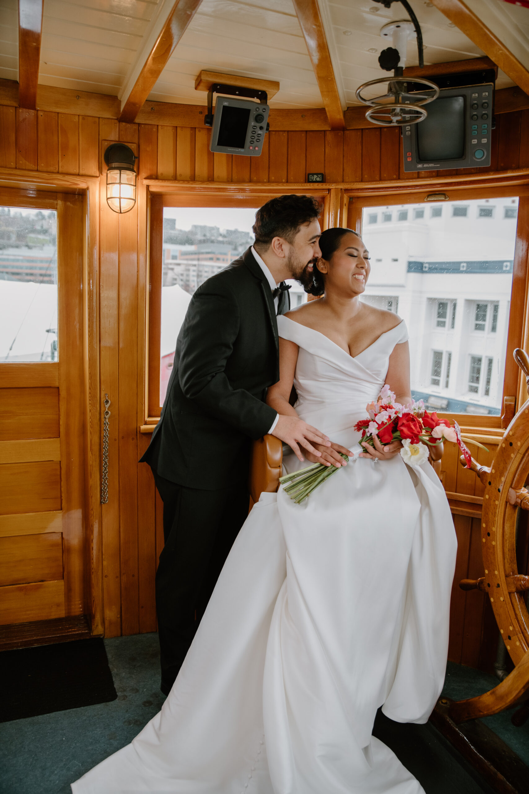 Groom kisses his bride as she smiles, holding a colorful bouquet inside the charming wheelhouse during their intimate boat wedding celebration.