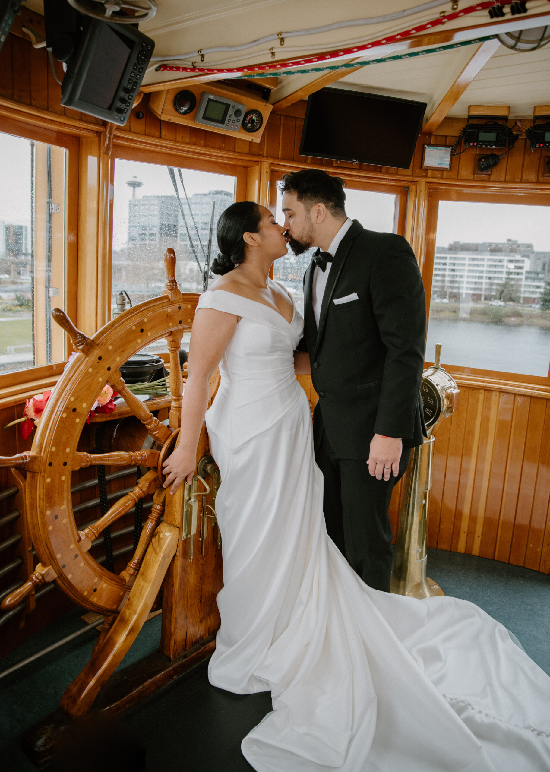 Bride and groom share a kiss beside the massive ship wheel, with city views behind them, showcasing the romance of a Virginia boat wedding venue.
