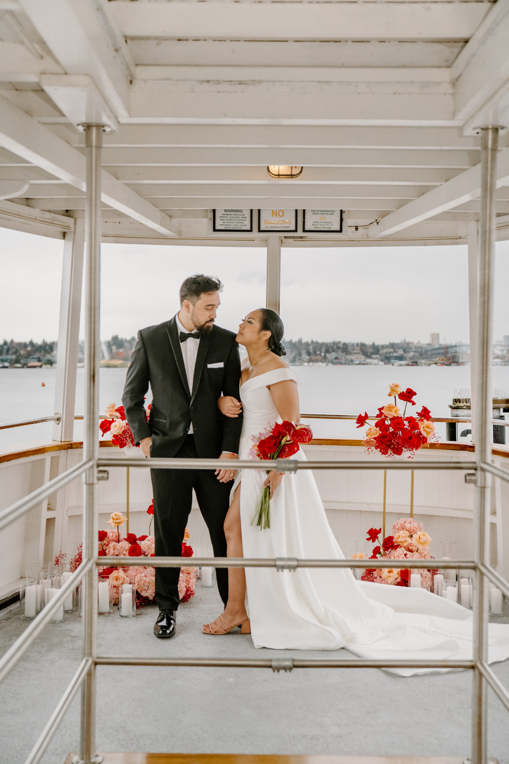 Bride and groom standing on the back deck of the boat, surrounded by romantic red floral arrangements at their boat wedding.