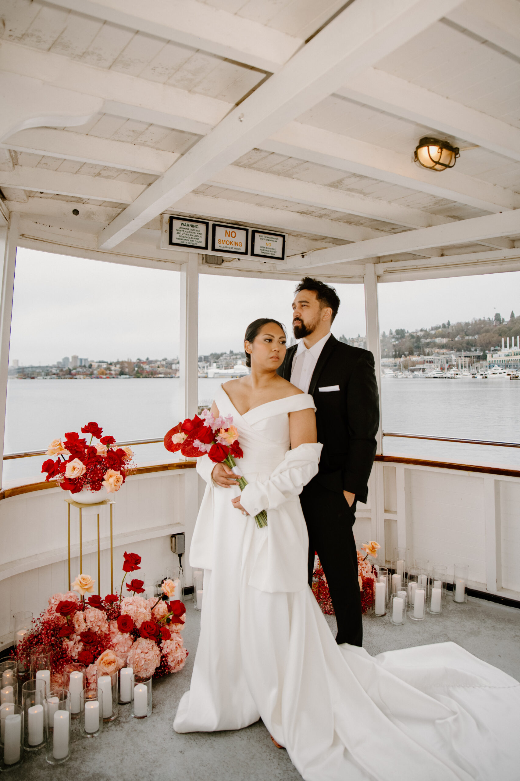 Bride and groom standing on the back deck of the boat, surrounded by romantic red floral arrangements at their boat wedding.
