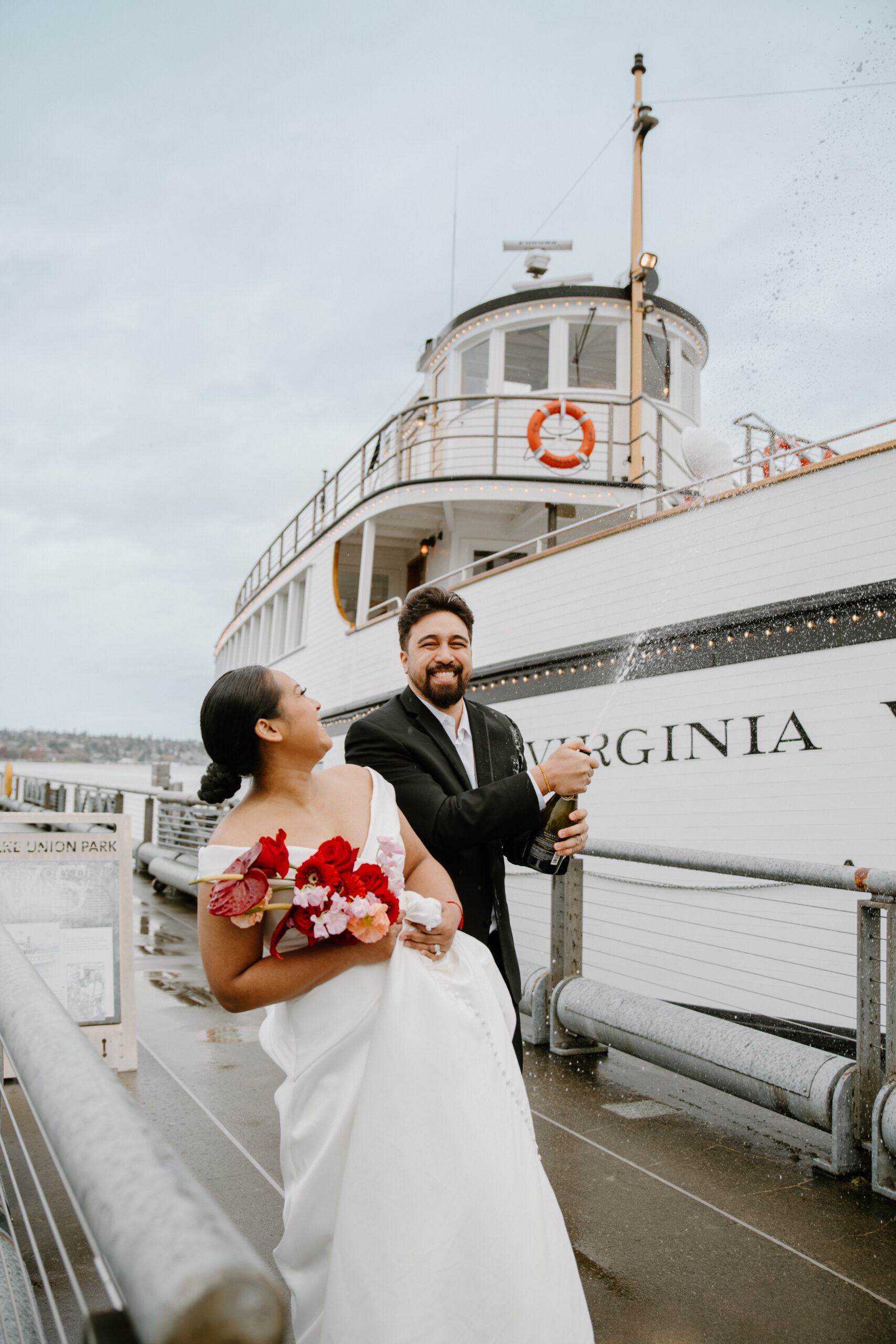 Groom popping champagne on the dock next to the historic Virginia wedding venue boat, bride laughing beside him.