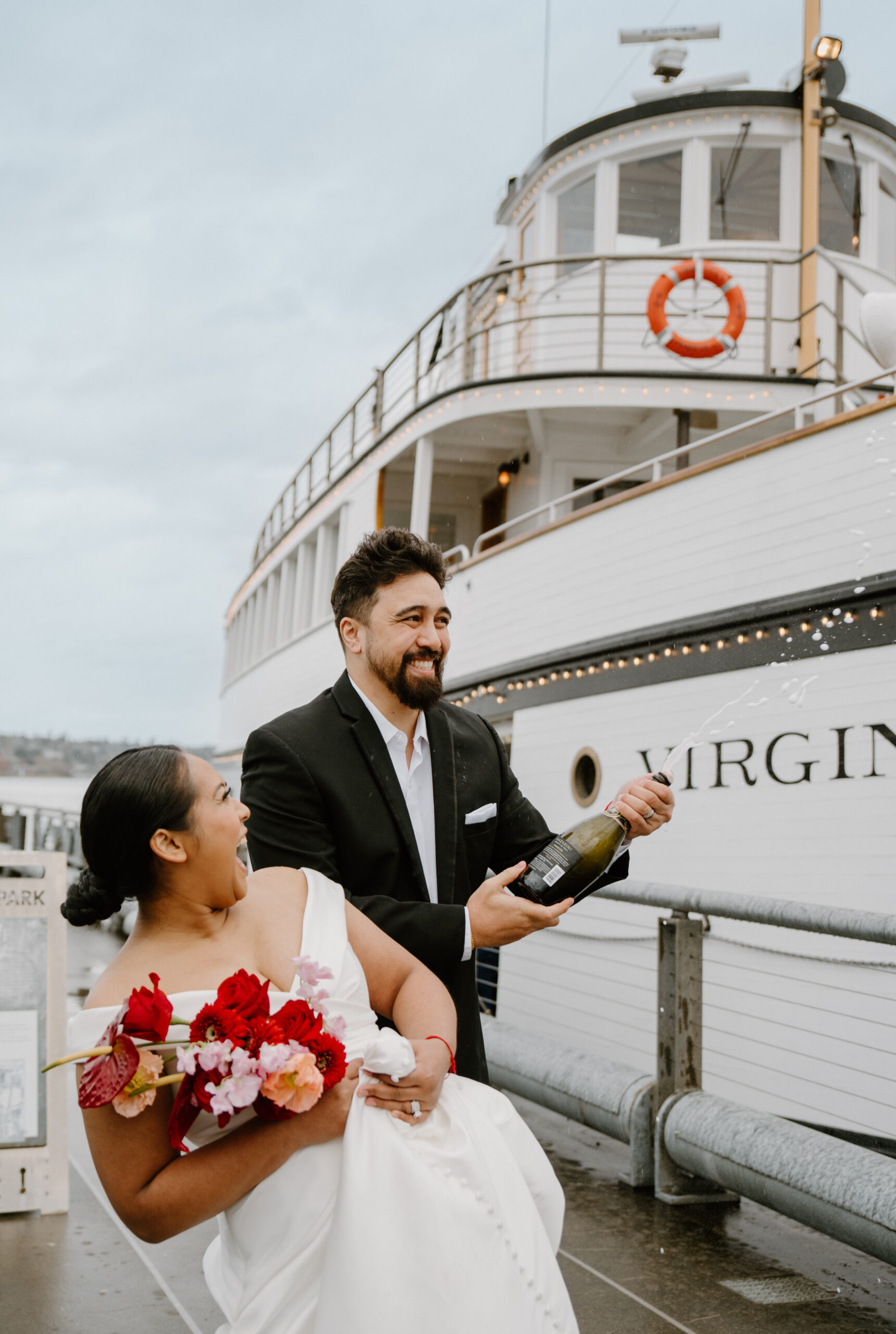 Groom popping champagne on the dock next to the historic Virginia wedding venue boat, bride laughing beside him.