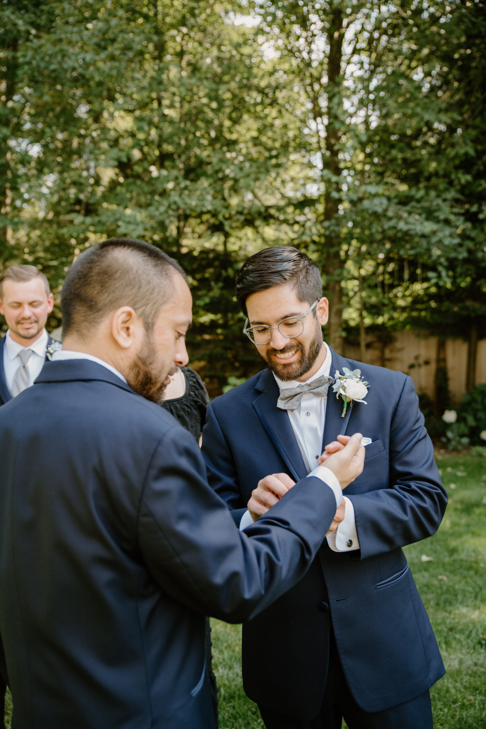Groom smiling as a friend pins his boutonnière, surrounded by his groomsmen in navy suits.