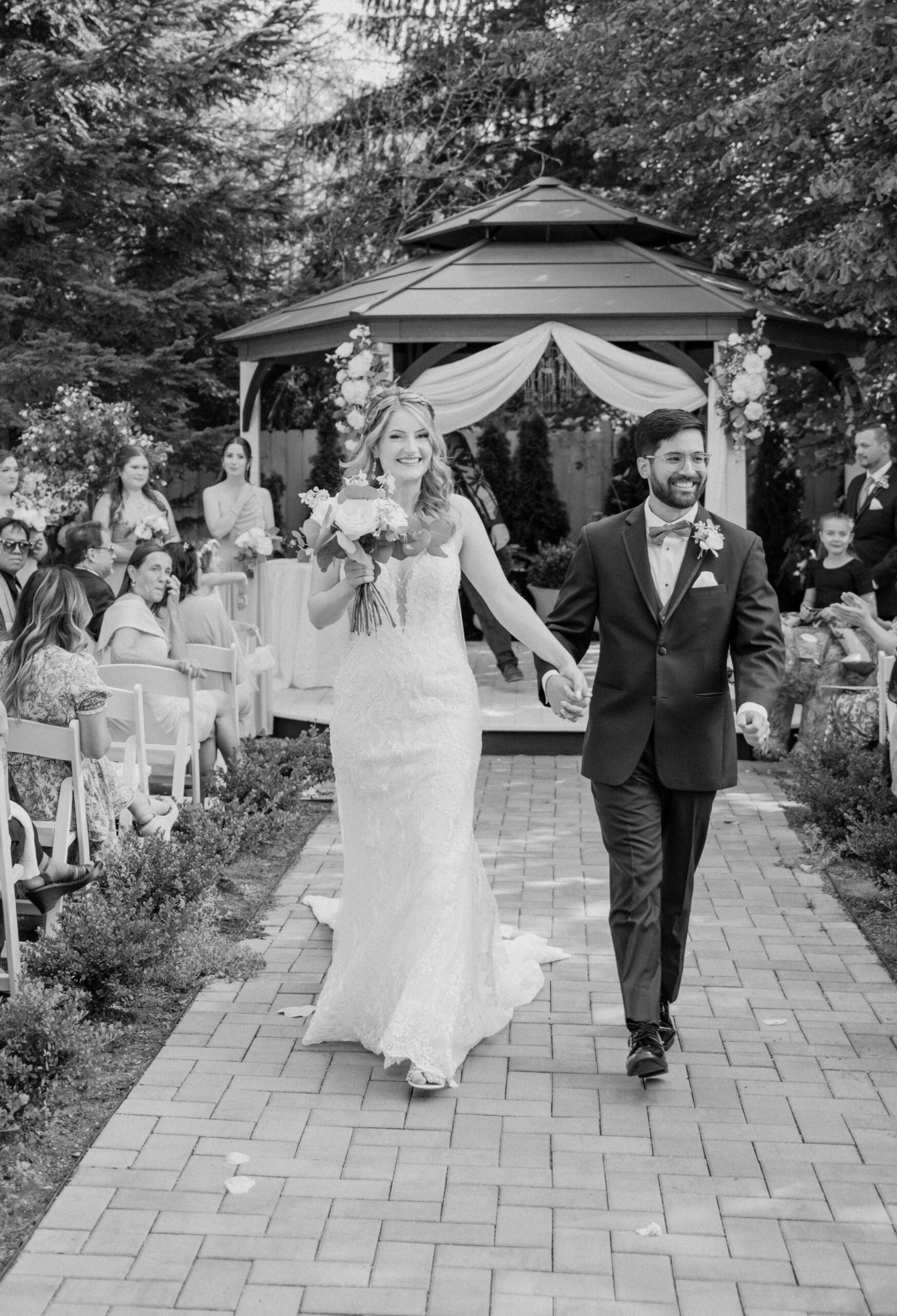 The newly married couple walks down the aisle beaming, surrounded by guests and lush greenery at Jackson Manor Events.
