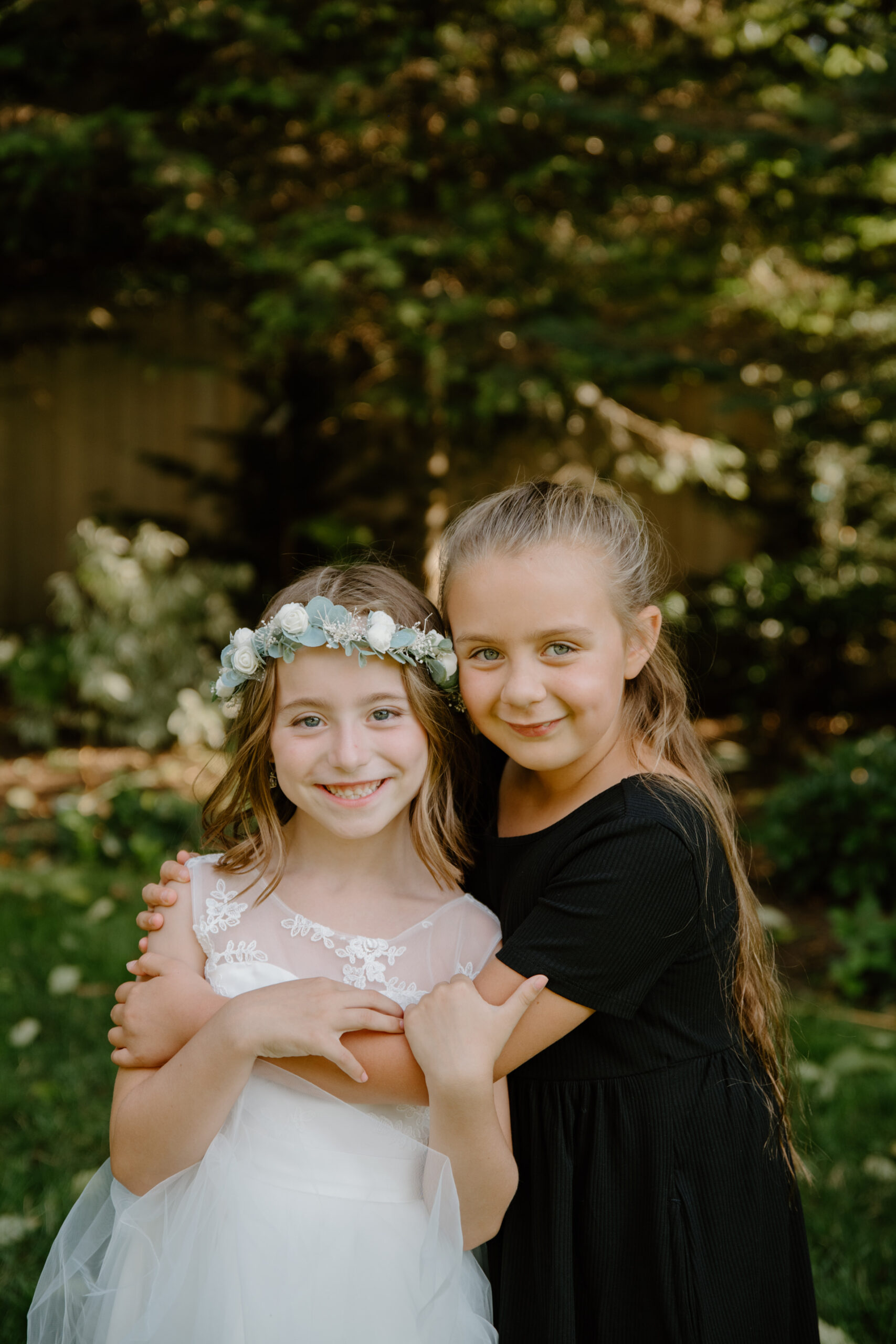 Two young flower girls smile sweetly while hugging, one wearing a white dress with a floral crown and the other in a black dress.
