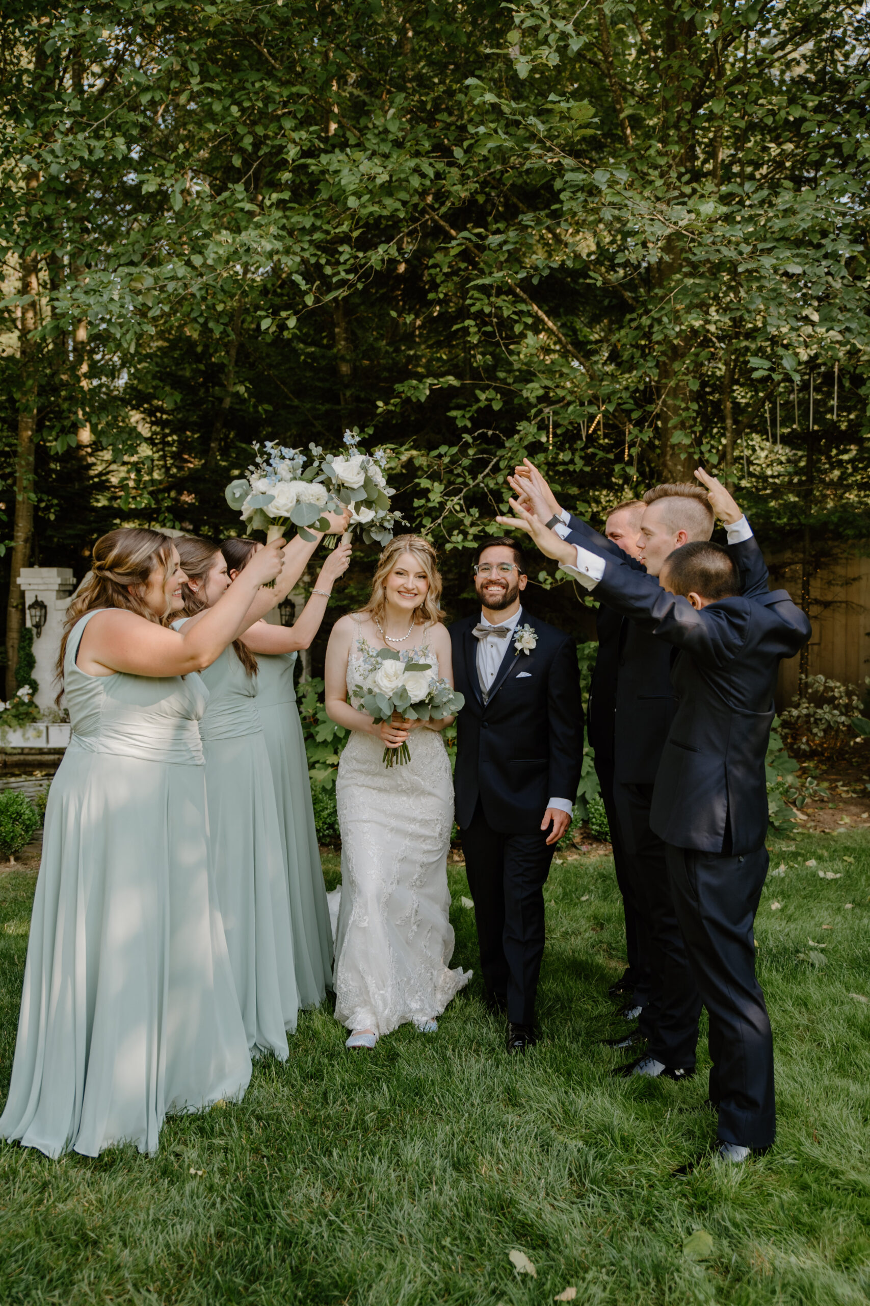 The wedding party raises their arms in celebration as the bride and groom walk together on the lawn, surrounded by trees at Jackson Manor Events.