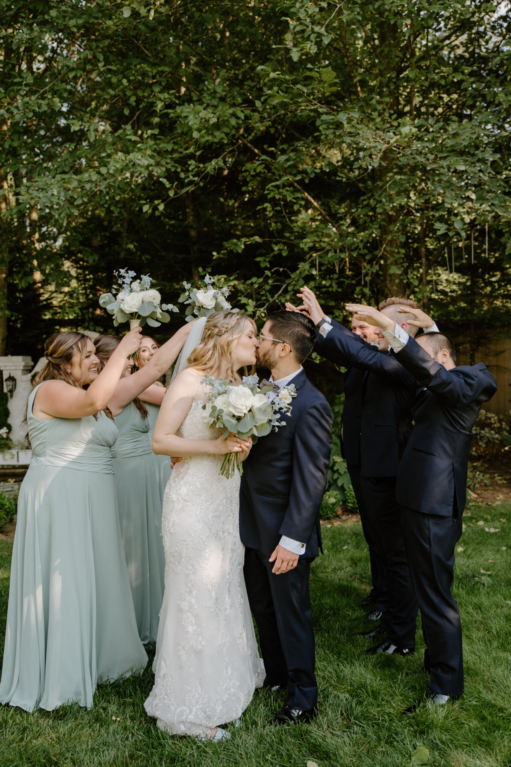 The bride and groom share a kiss while their wedding party cheers them on, holding floral bouquets in the garden.