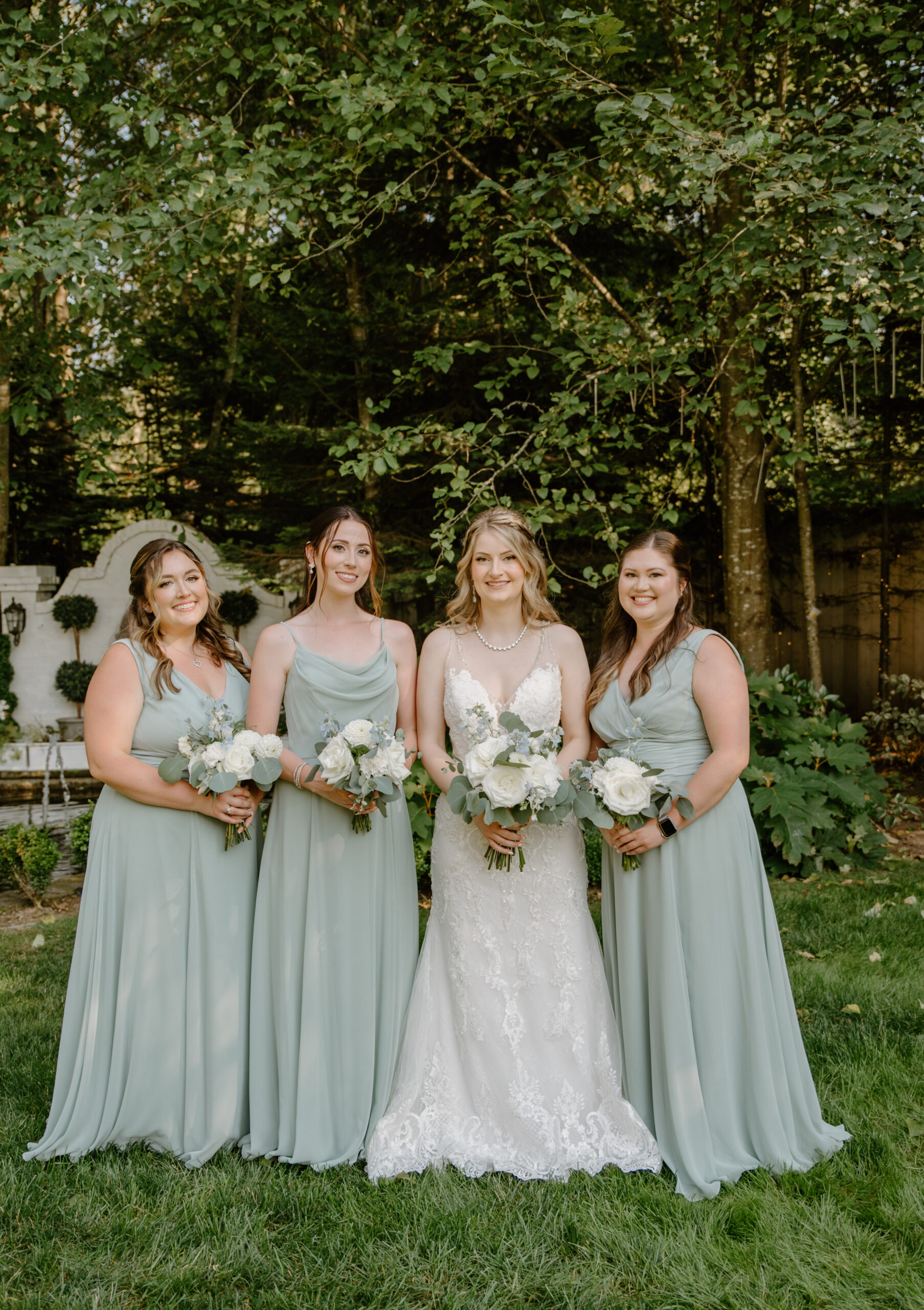 The bride poses with her three bridesmaids in soft green dresses, holding white bouquets in the garden at Jackson Manor Events.