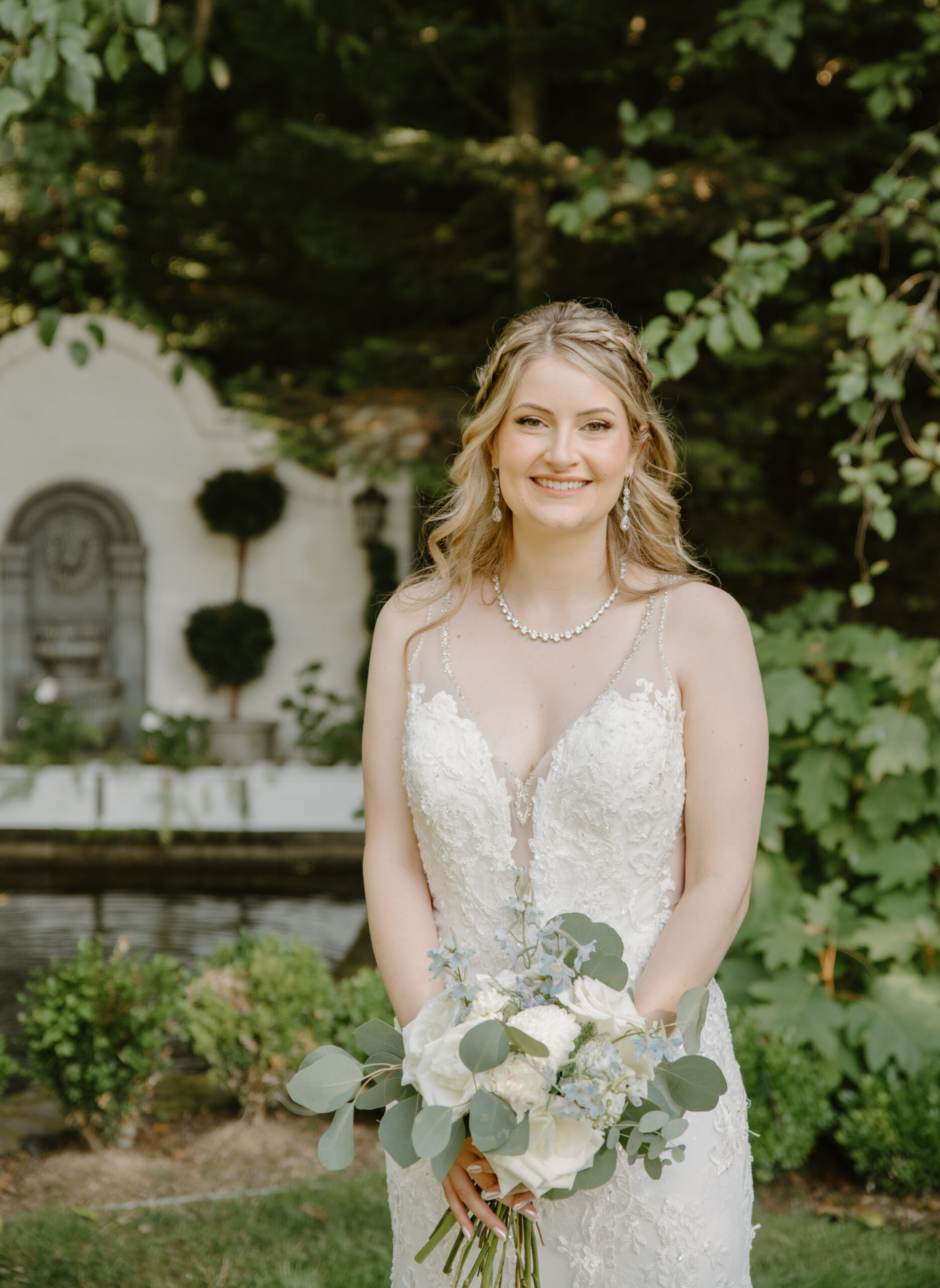 Bride stands with her bouquet of white roses and soft blue florals in front of the garden fountain at Jackson Manor Events, wearing a detailed lace gown.