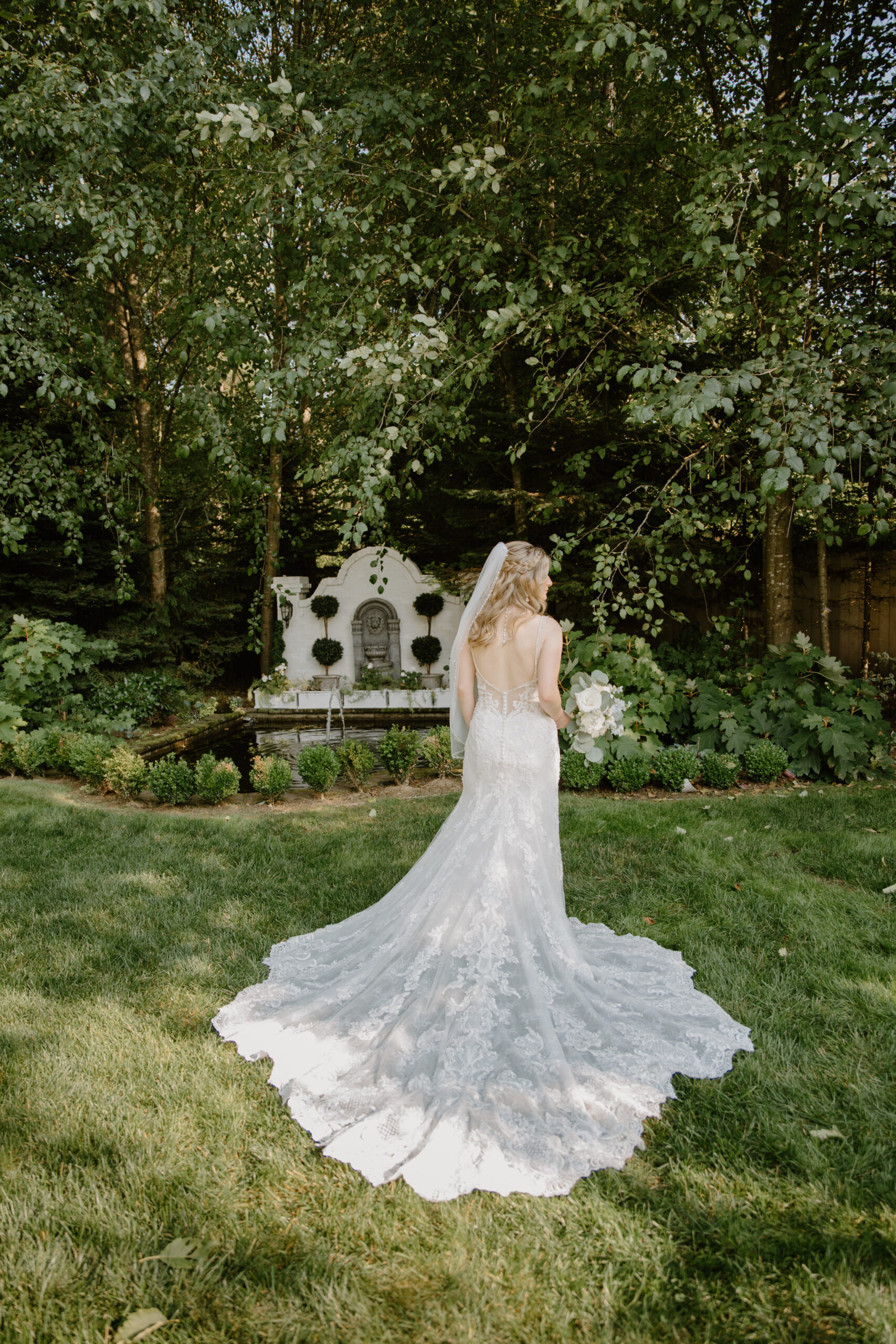 Back view of the bride walking toward the fountain at Jackson Manor Events, her long lace train trailing behind her across the lawn.