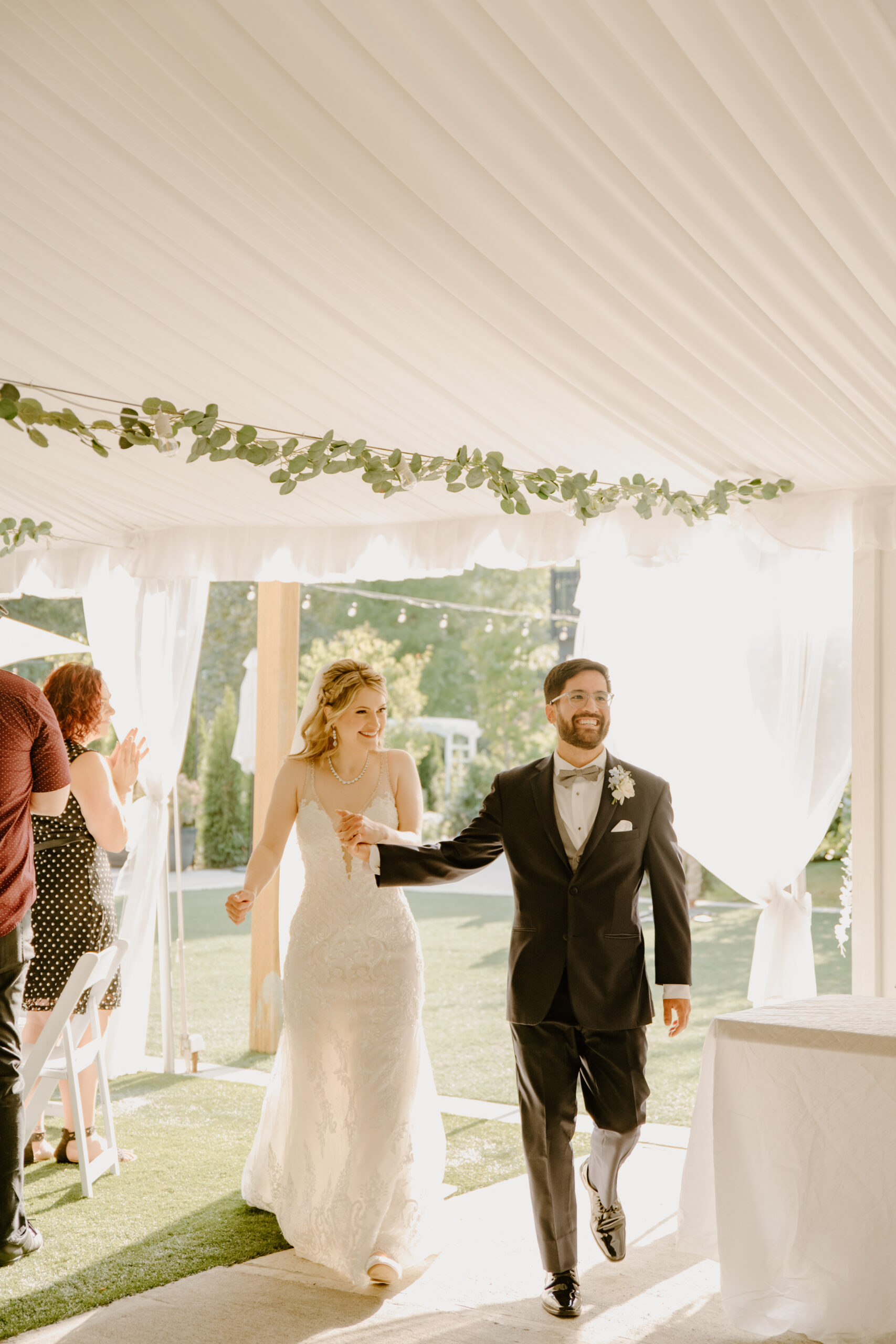 Bride and groom make their grand entrance into the tented reception area, holding hands with joyful expressions as guests applaud.