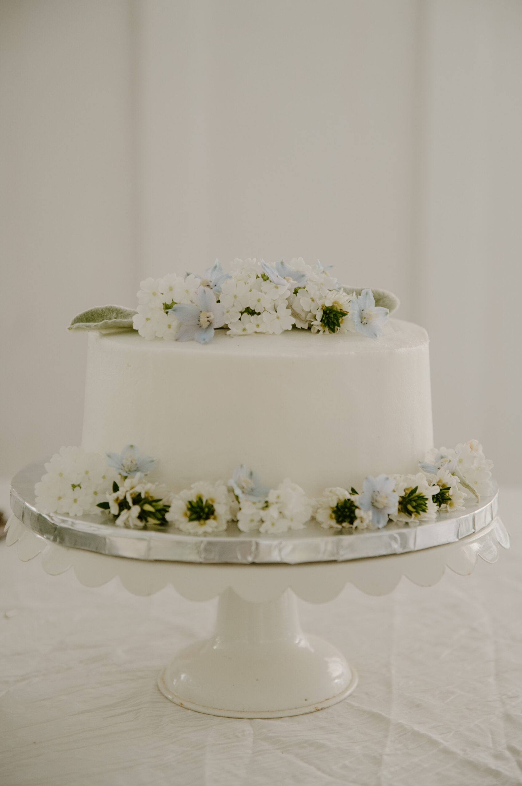 Simple white wedding cake adorned with white and pale blue flowers, sitting on a scalloped cake stand against a neutral backdrop.
