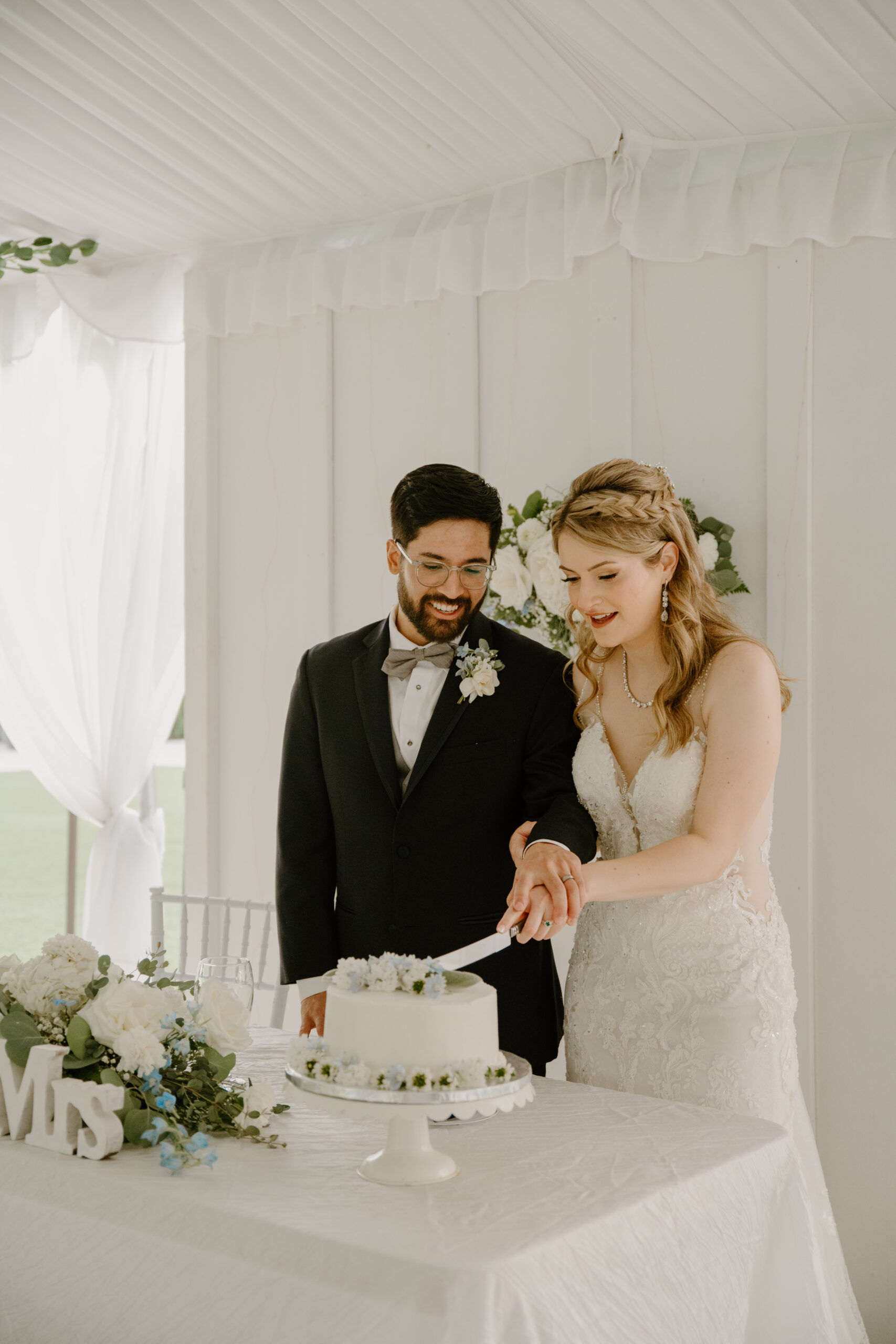 Bride and groom cut their wedding cake together under the reception tent, smiling and enjoying the moment with their guests nearby.