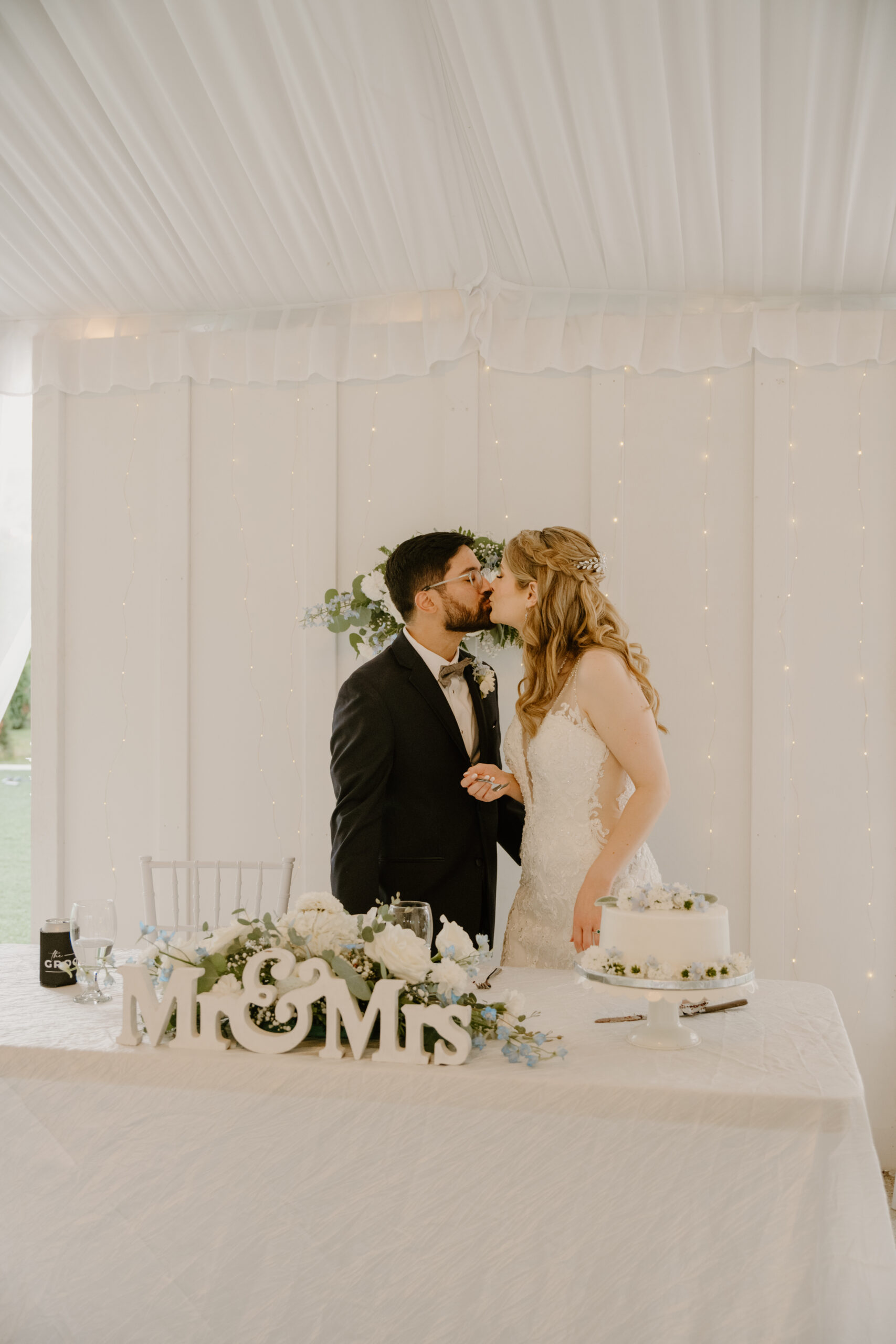 The bride and groom share a kiss behind their sweetheart table adorned with a “Mr & Mrs” sign, fresh floral arrangements, and their white wedding cake at Jackson Manor Events.