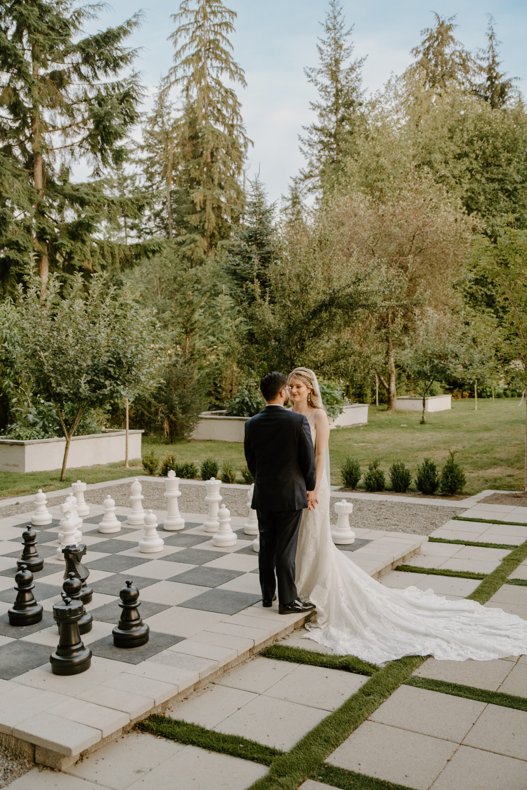 The groom gazes at his bride as they stand together at the edge of the giant chessboard surrounded by trees and evening light.
