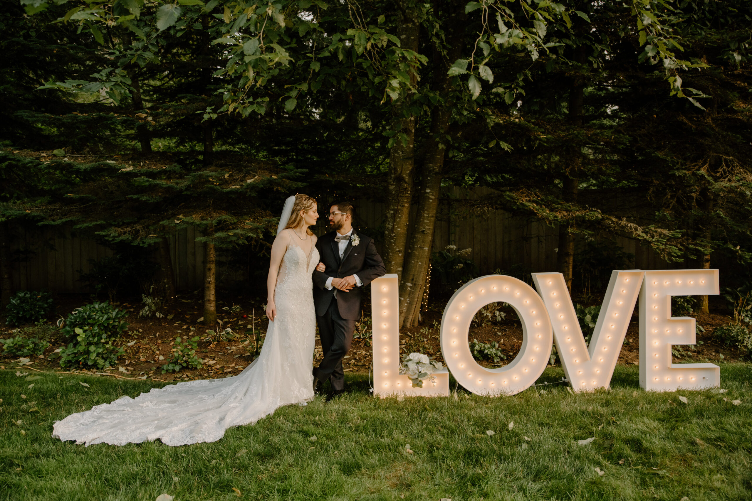The bride and groom stand beside an illuminated LOVE sign in front of towering trees, adding a whimsical touch to their Jackson Manor Events celebration.