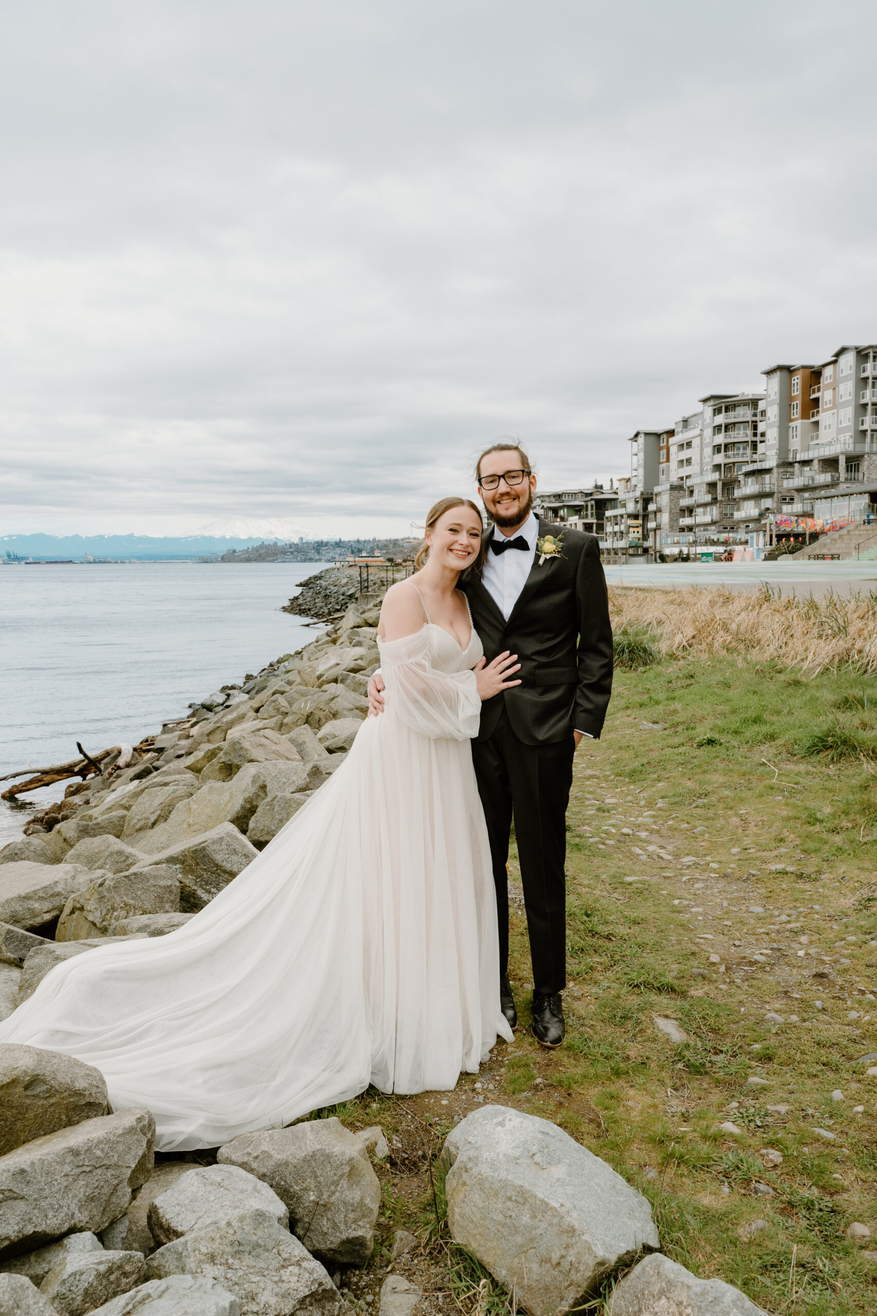 A joyful bride and groom pose together along a rocky shoreline with city buildings in the background, celebrating their waterfront PNW wedding.