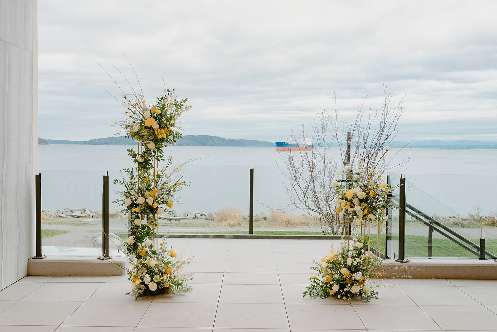PNW wedding ceremony arch with yellow and white florals framing a serene water view under an overcast sky. 