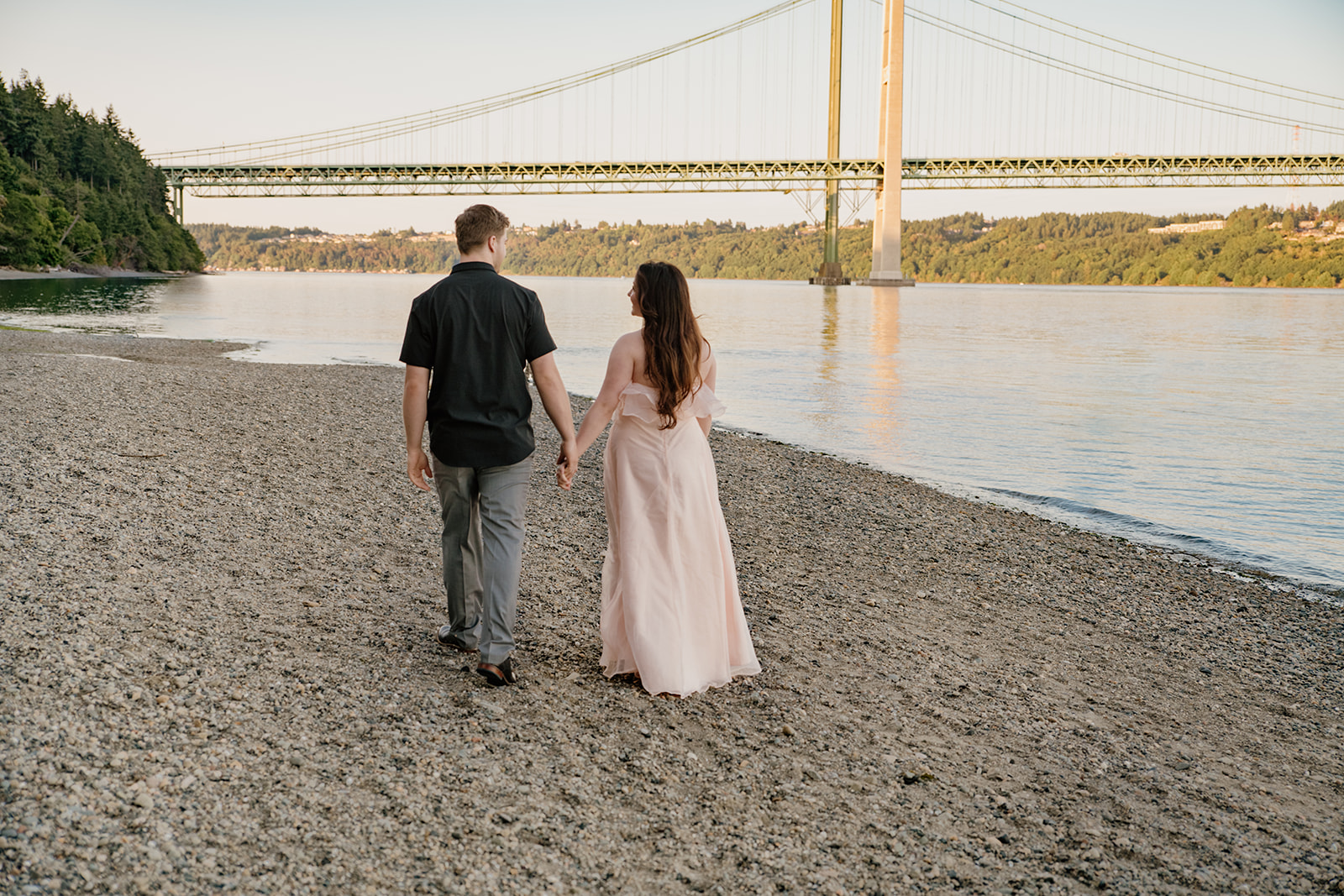 Couple walking hand in hand on a rocky beach with a bridge in the background, one of the more unique waterfront engagement photo locations.
