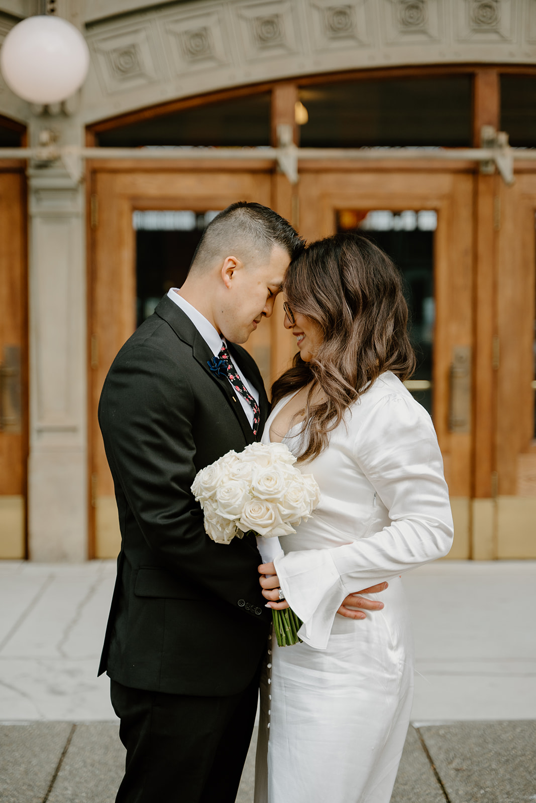 Bride and groom stand forehead to forehead outside the Tacoma courthouse, holding white roses after their courthouse elopement.