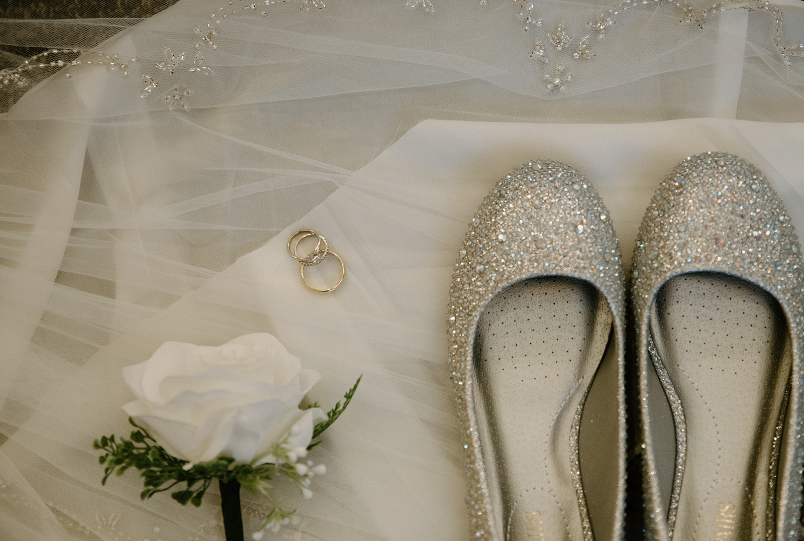 Flat lay of glittery wedding shoes, rings, and boutonnière on bridal veil.