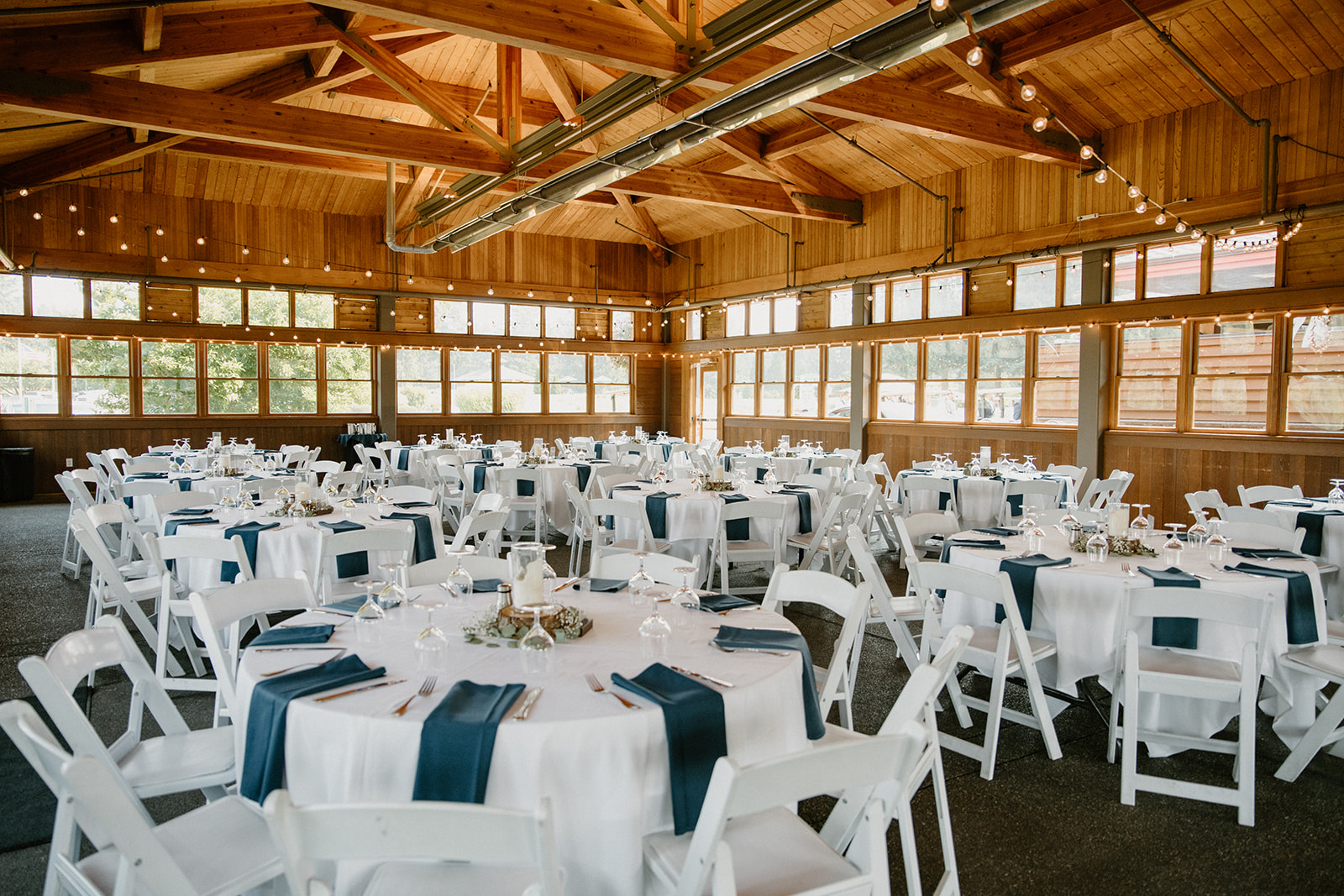Wide shot of the wedding reception setup with round tables, navy linens, and string lights inside a wooden venue.
