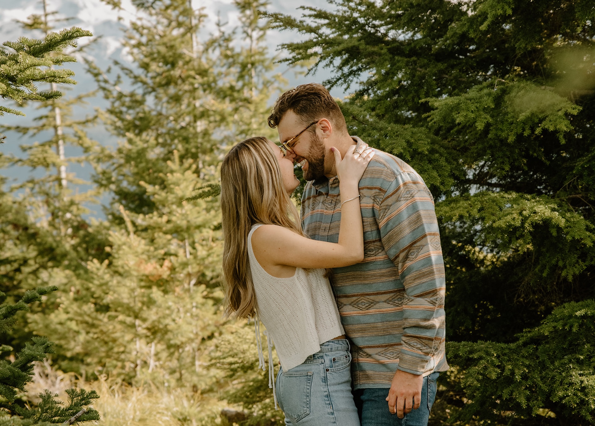 A playful and candid engagement shoot moment with a couple laughing and leaning into each other among tall evergreens. 
