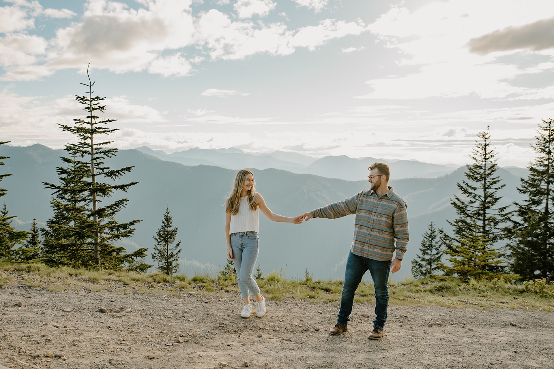 The couple holding hands while walking along a mountain trail, framed by panoramic views of forested hills and dramatic clouds. 