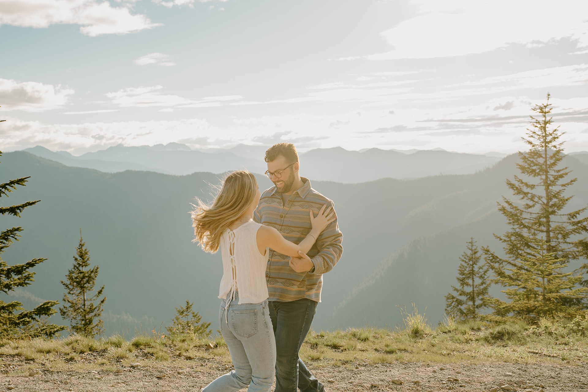 A playful couple dancing on a mountaintop with the golden sunset casting light across the mountain range behind them. 