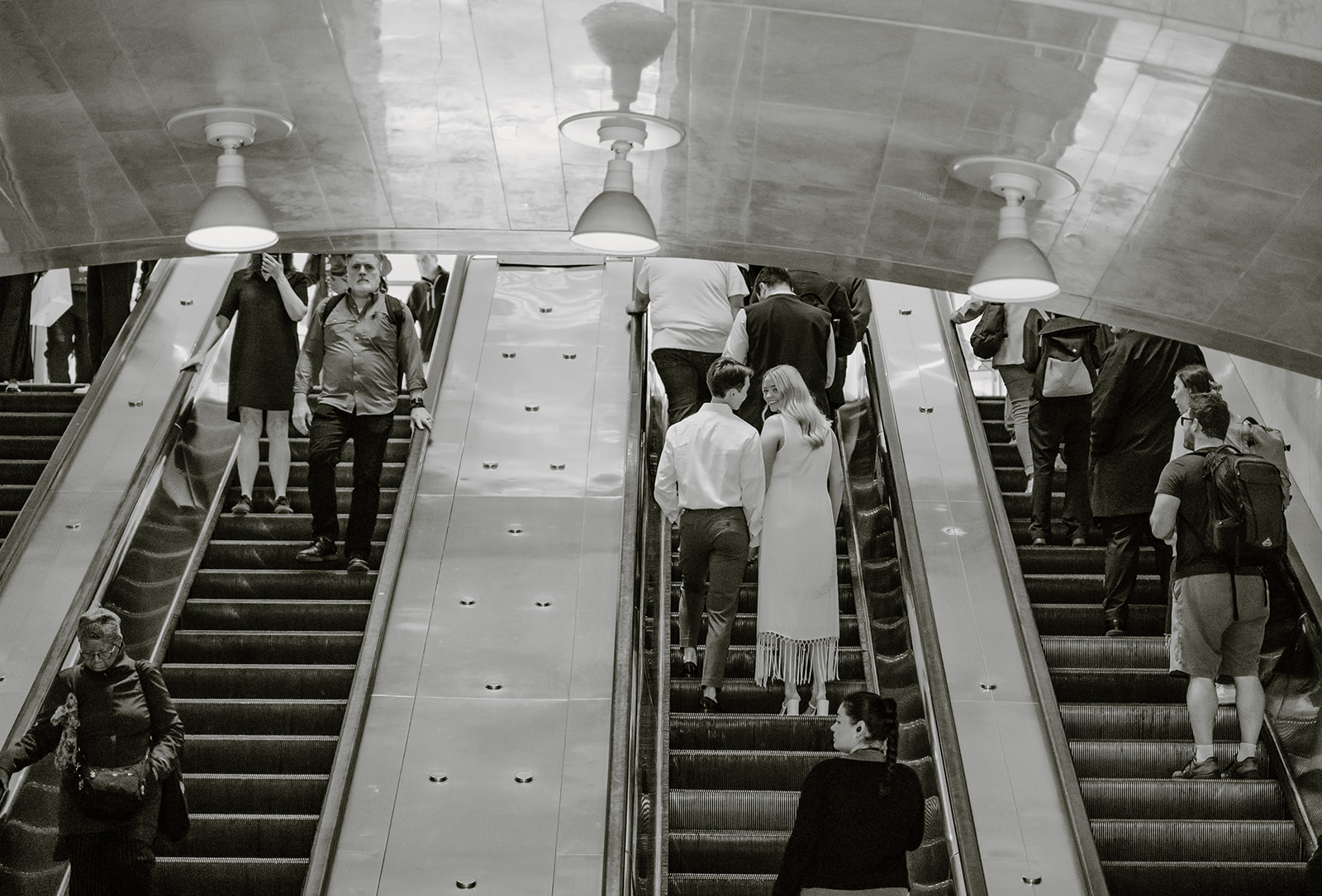 A black and white photo of the engaged couple holding hands going up the escalator at Grand Central Station during their New York City Engagement Photos