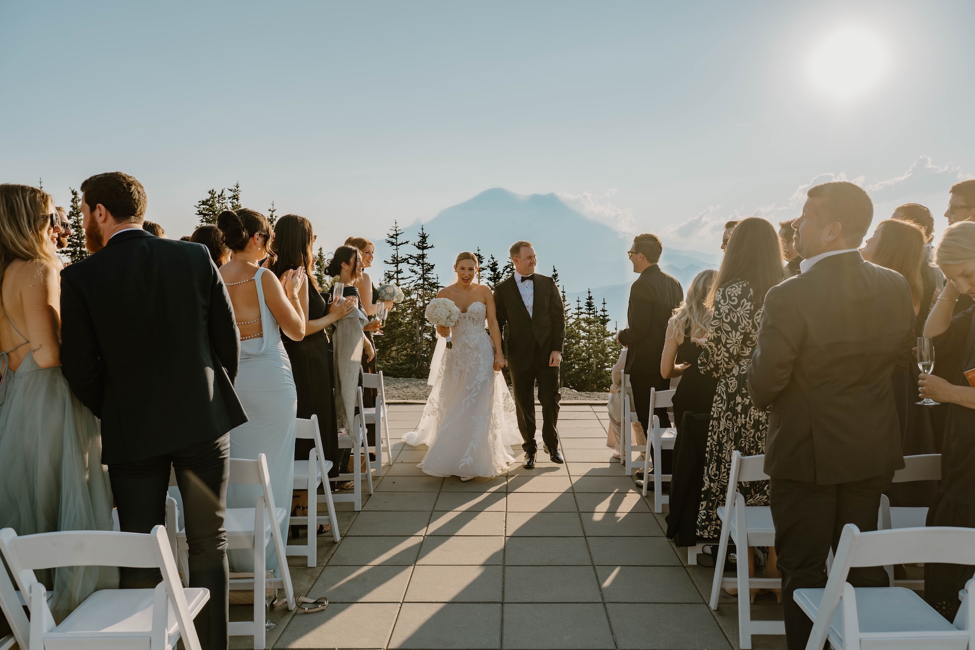 Bride and groom walking up the ceremony aisle outside with guests standing and clapping during a sunny mountain-top celebration.