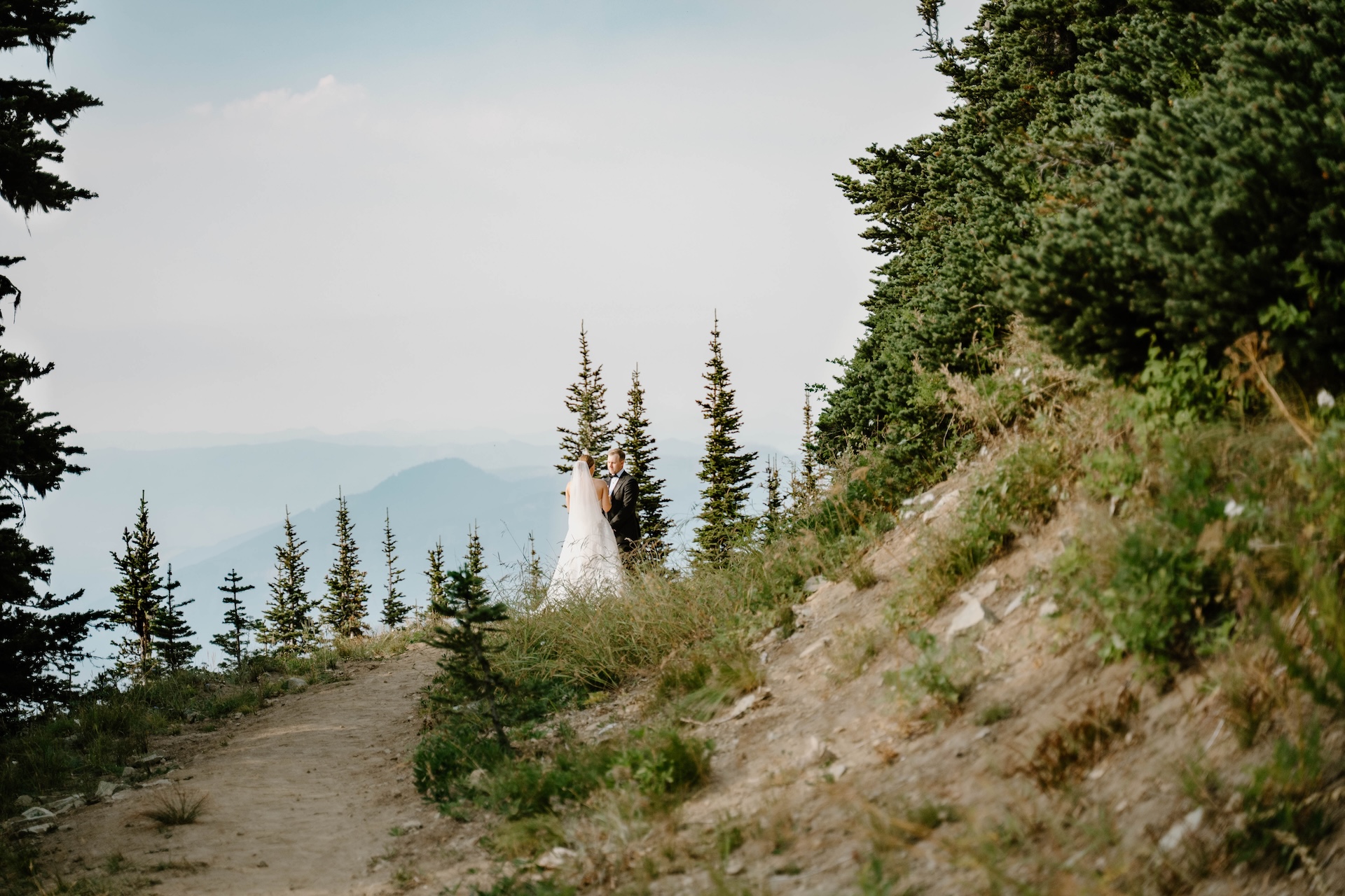 Couple standing on a mountain ridge trail with tall evergreens and hazy peaks in the background during a Crystal Mountain Resort Wedding.