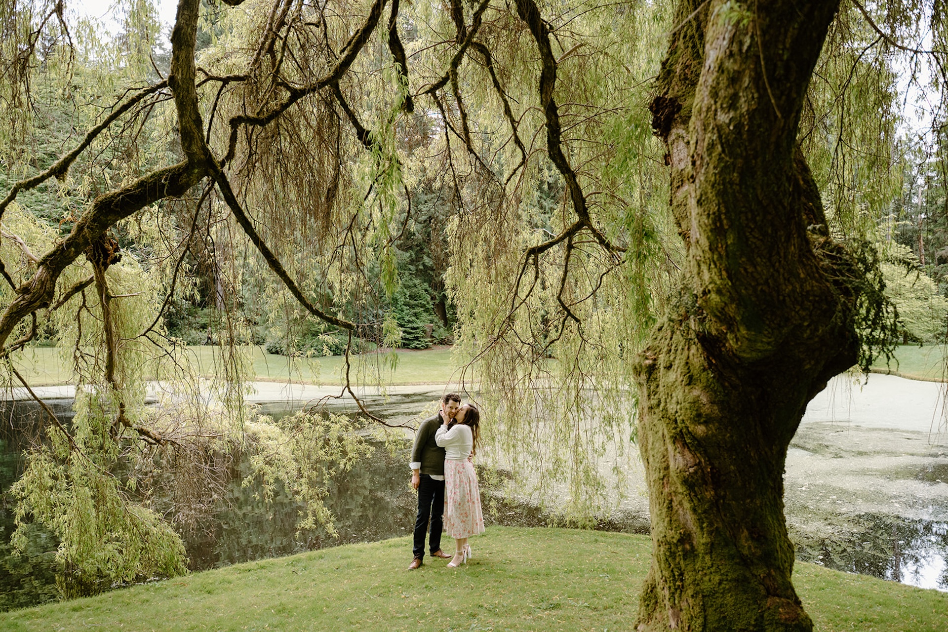 Woman kissing her fiances cheek while they stand under a willow tree overlooking a pond at Bloedel Reserve in Washington