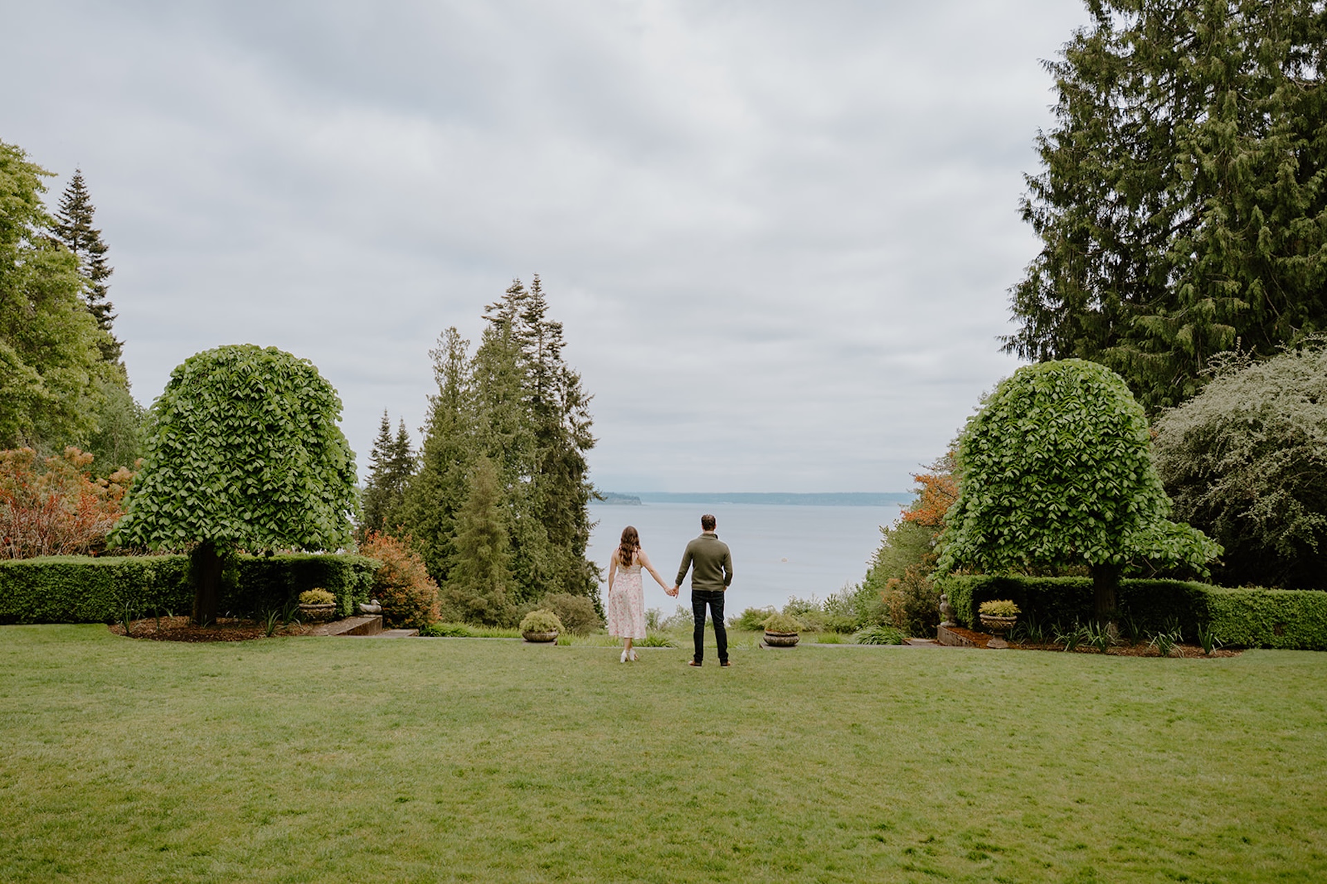 Couple holding hands in a lawn at Bloedel Reserve overlooking the water during their engagement shoot.