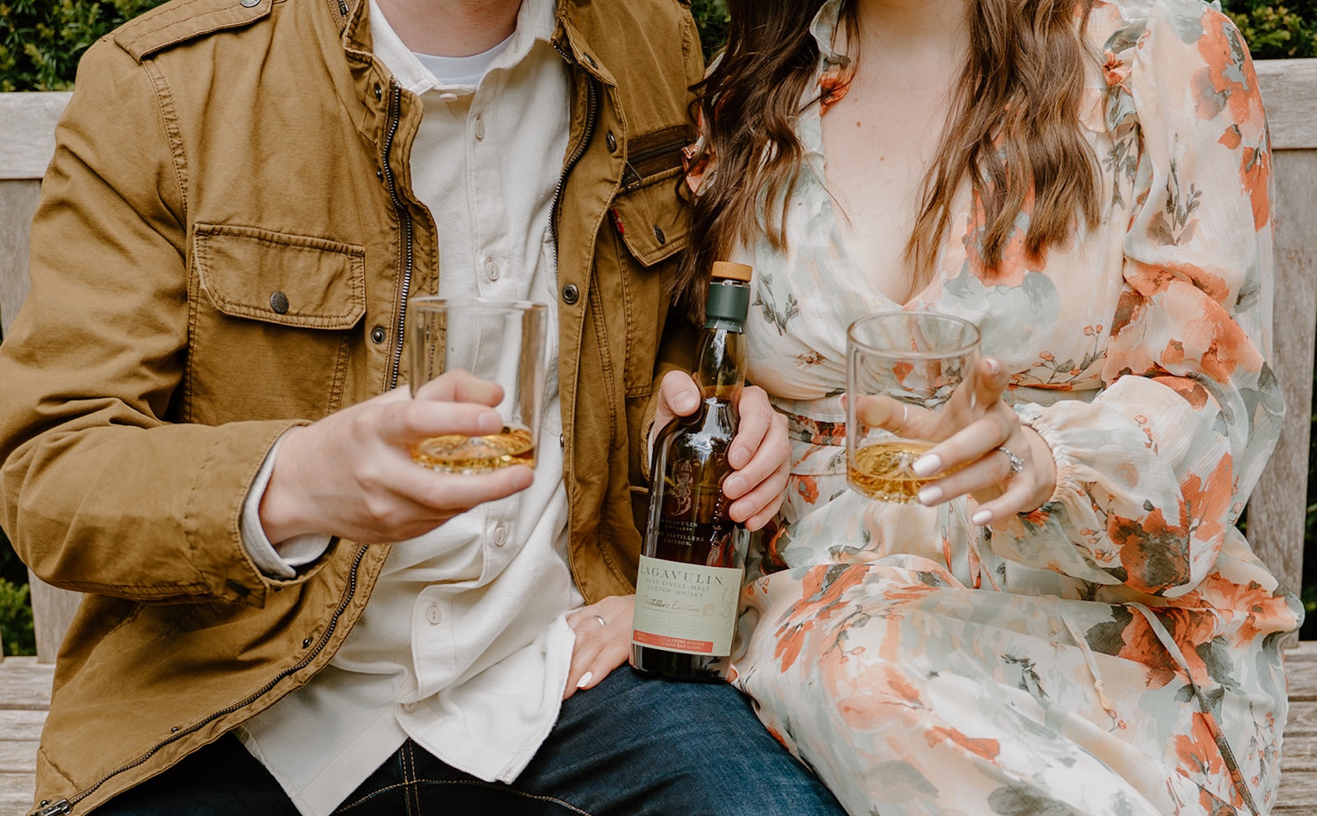 Couple holding glasses of whiskey during their engagement shoot while sitting on a park bench