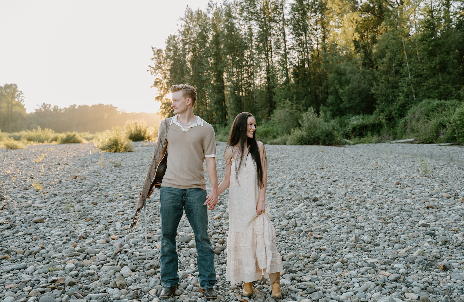Couple holding hands and smiling while looking away from each other during their golden hour portraits on a bed of rocks by a river with the sunset behind them.