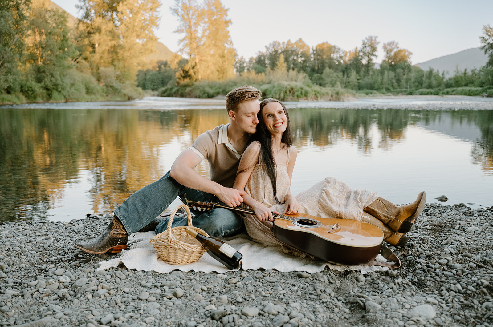 They sit on a blanket by the river with a guitar and picnic basket, leaning into each other and showcasing relaxed When to take engagement photos moments.