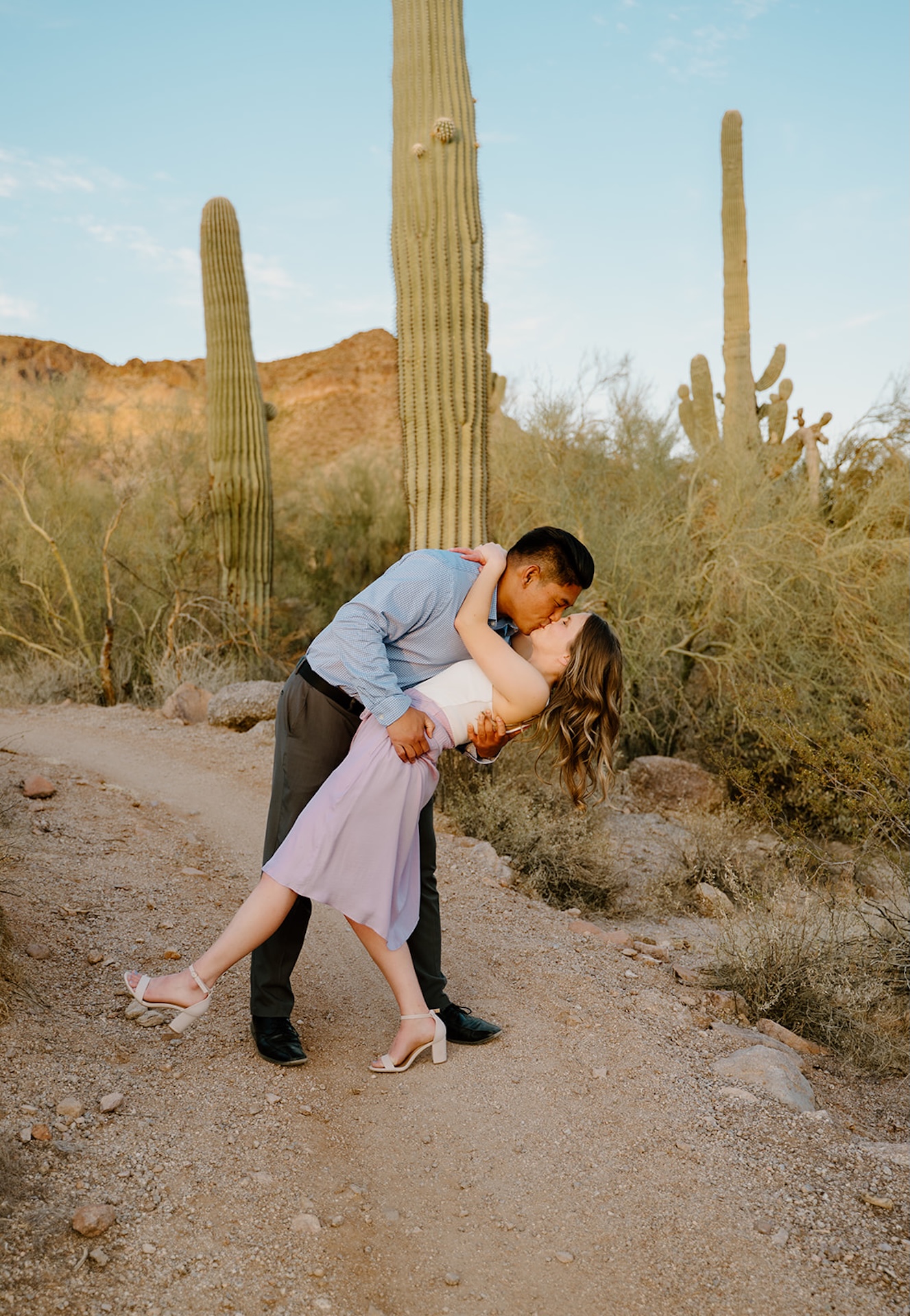The groom dipping the bride playfully on a dirt path with tall cacti and desert greenery framing the scene.