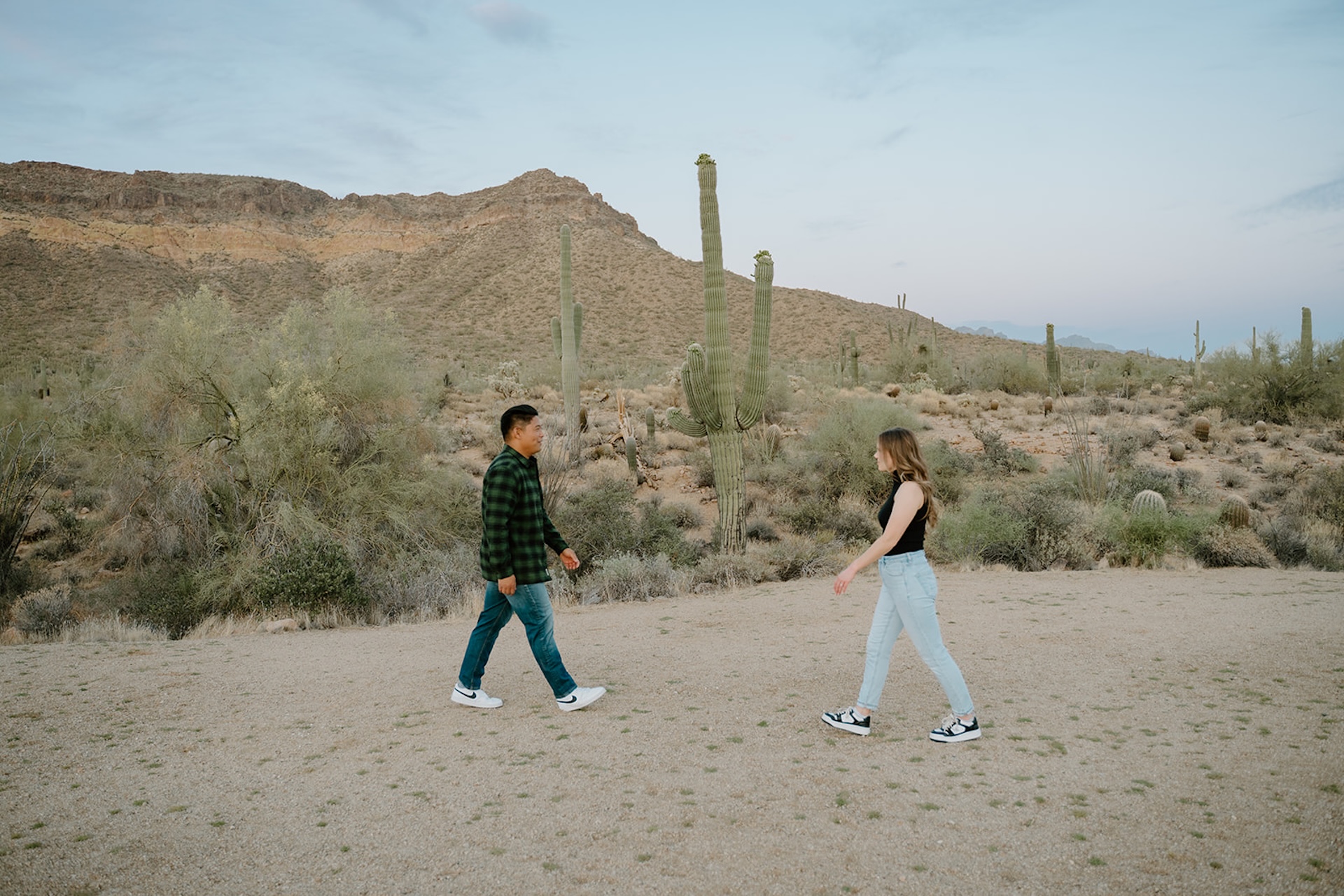 A wide shot of the couple walking toward each other on a dirt path with towering saguaro cacti and mountains behind them, captured as part of arizona engagement photos.
