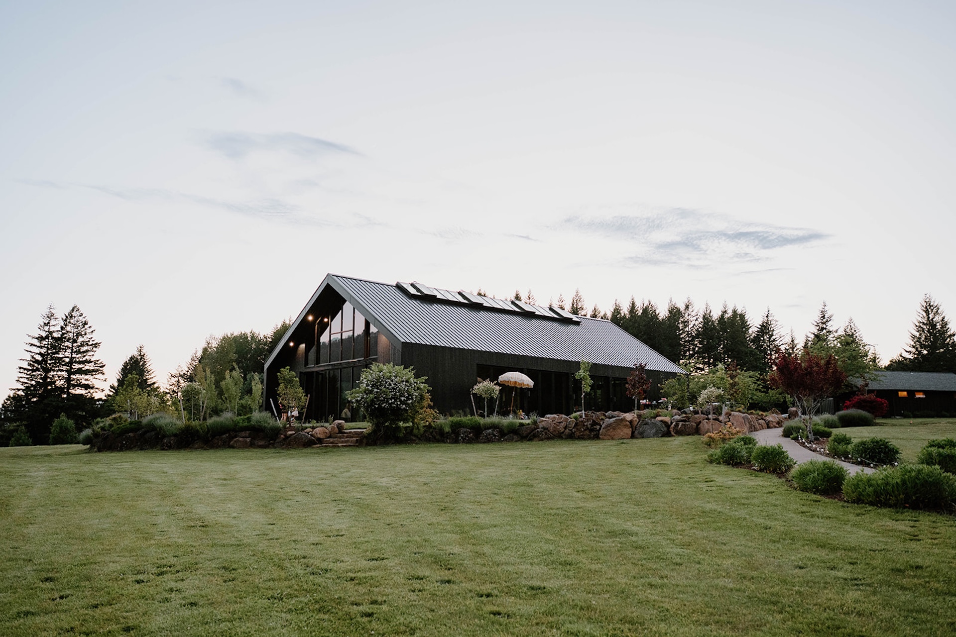 Wide exterior view of the modern black barn venue surrounded by green lawn and trees during a wind mountain ranch wedding, captured in soft evening light.