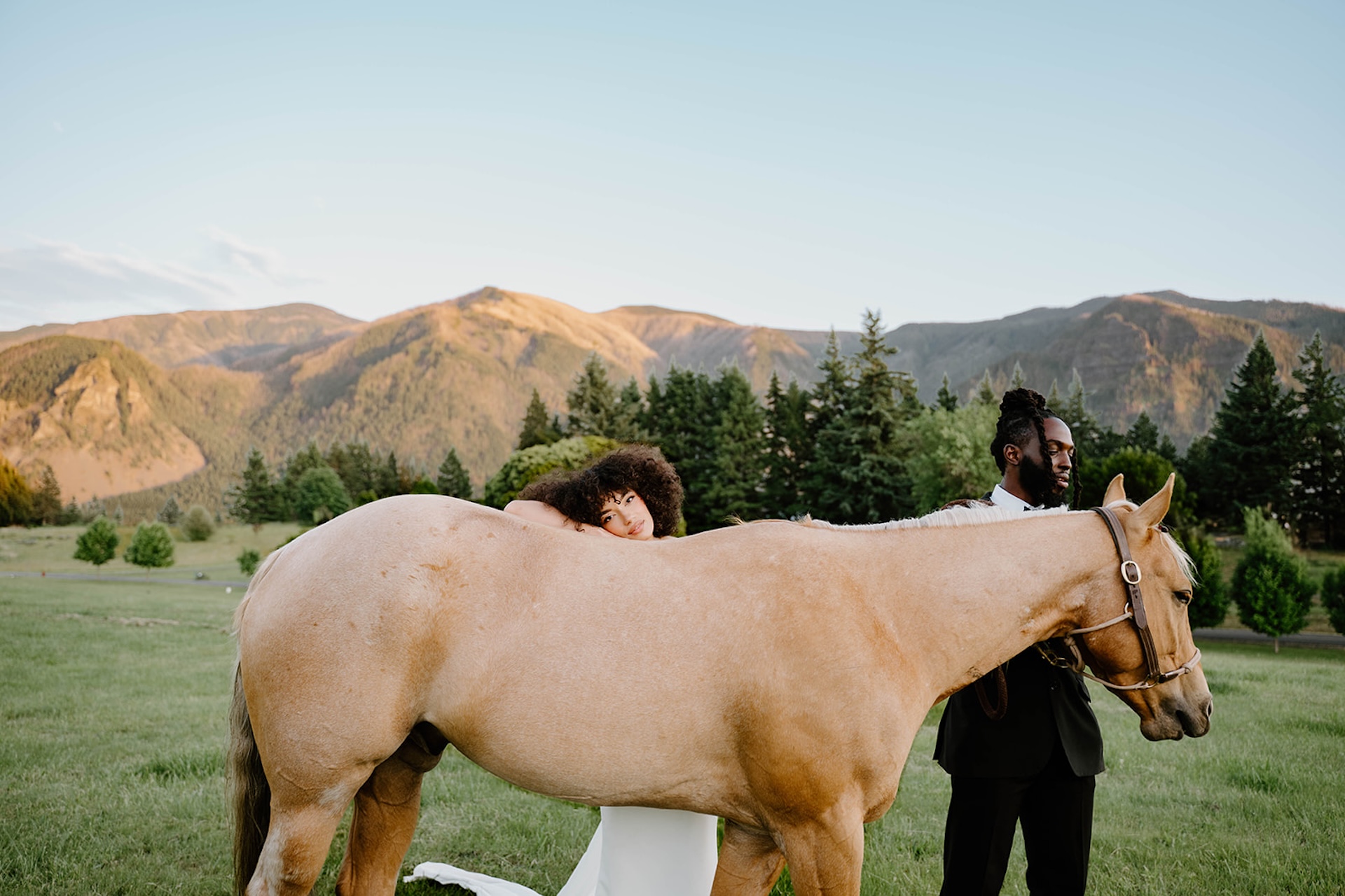 Color portrait of the bride resting against a horse while the groom stands nearby, showcasing editorial styling, natural light, and sweeping mountain scenery.