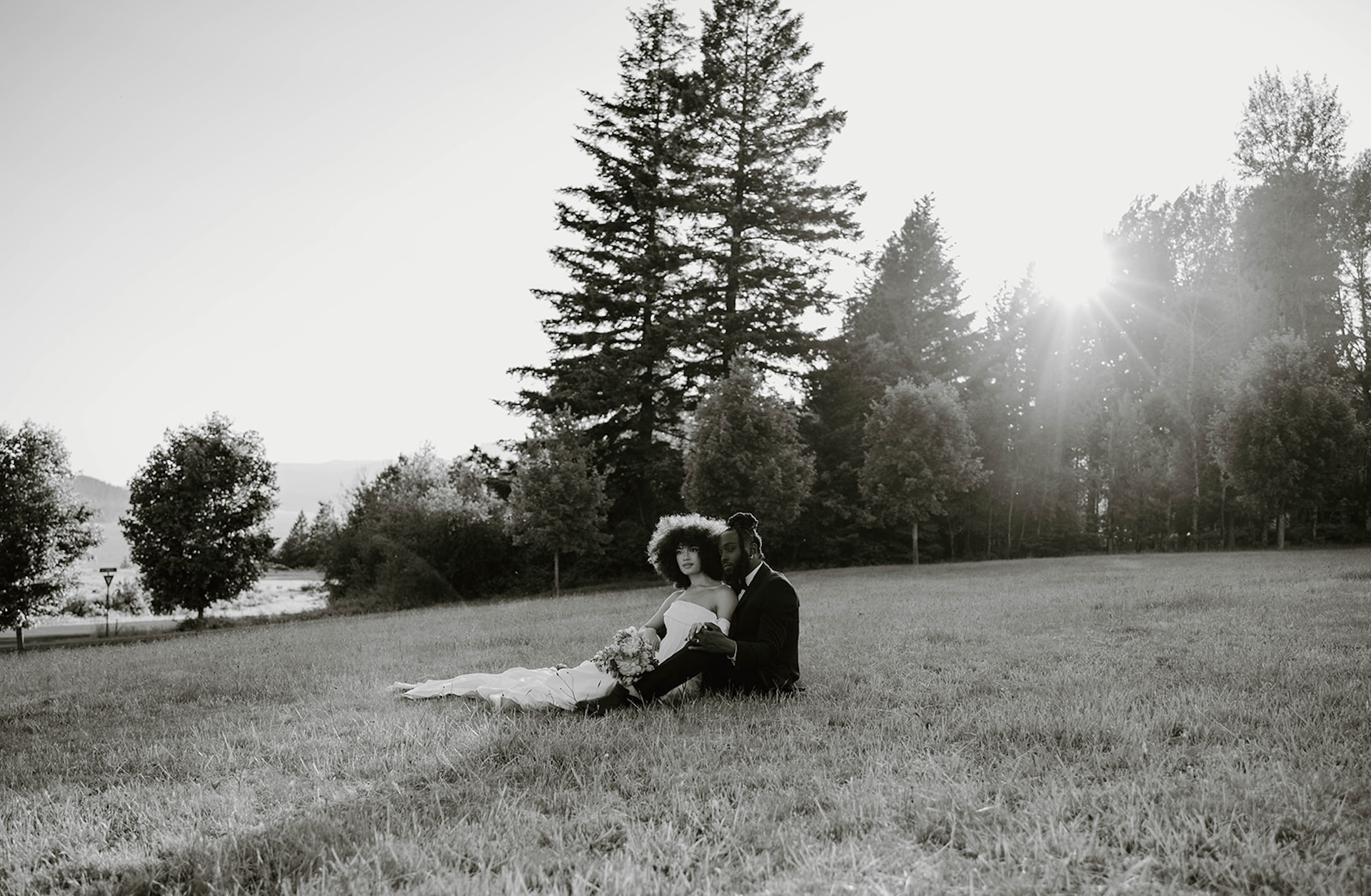 Black and white portrait of the couple sitting together in an open grassy field, with trees and sunlight creating a calm, intimate mood.