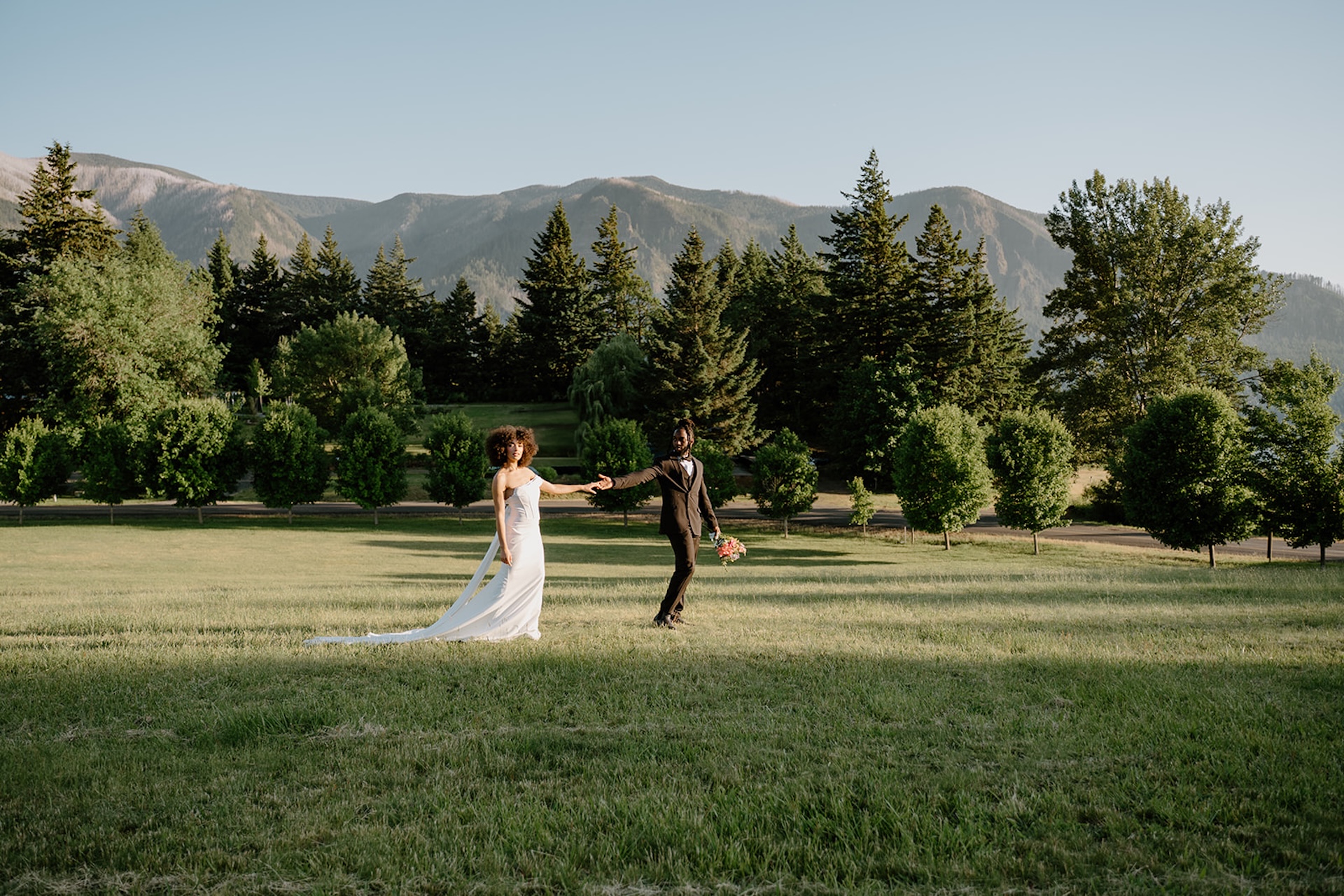 Wide portrait of the bride and groom holding hands while walking across an open field with mountain views and rows of trees in the background.