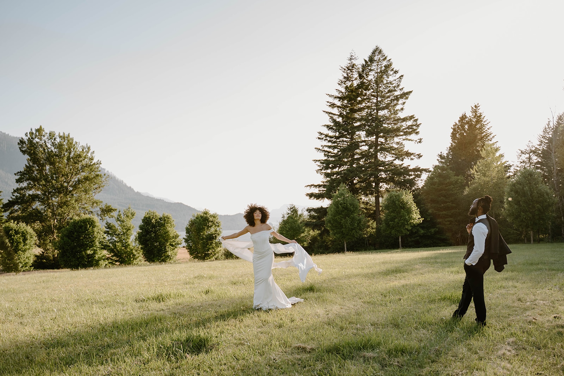 Wide scenic portrait of the bride and groom standing apart in an open meadow, showcasing flowing fabric, tall trees, and expansive mountain views.