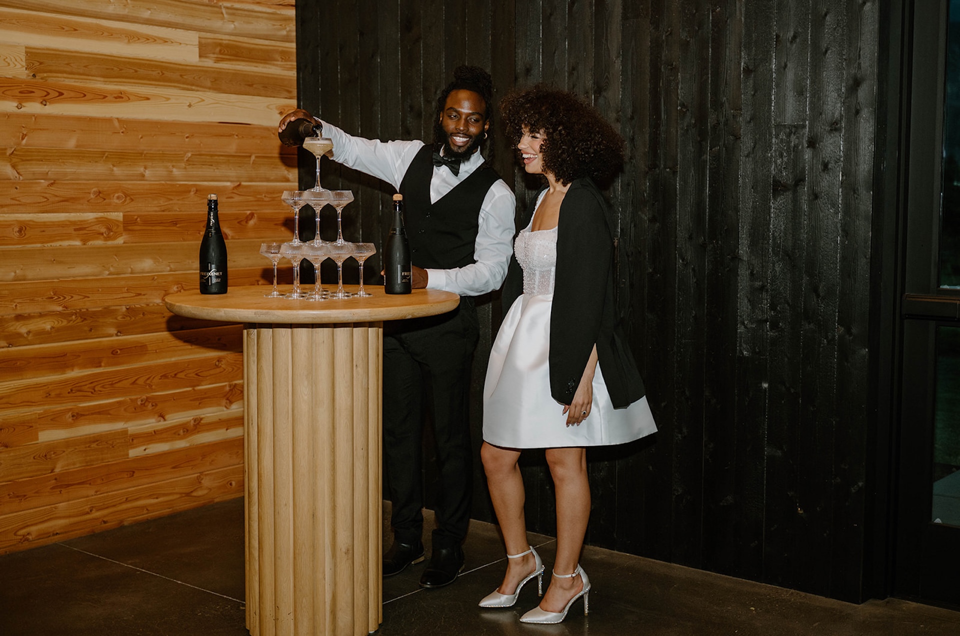 Bride and groom laughing together indoors while pouring champagne into a stacked coupe tower against warm wood walls.