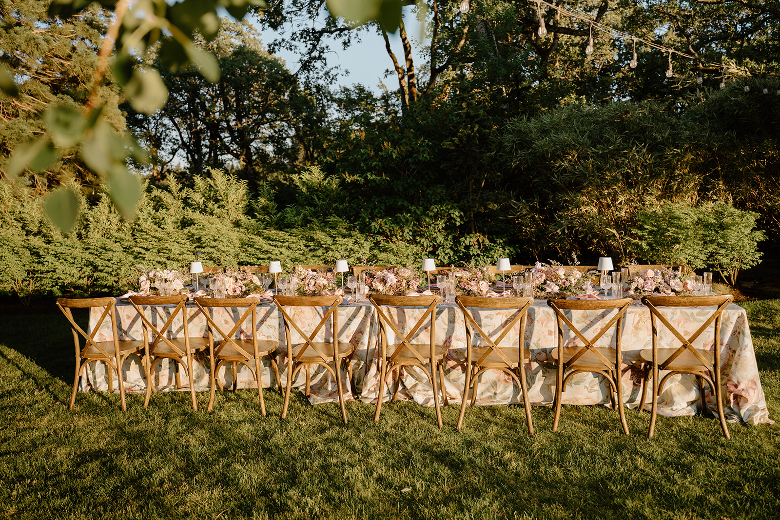 Long wooden reception table styled with floral tablecloths, blush napkins, and garden-inspired centerpieces under string lights.