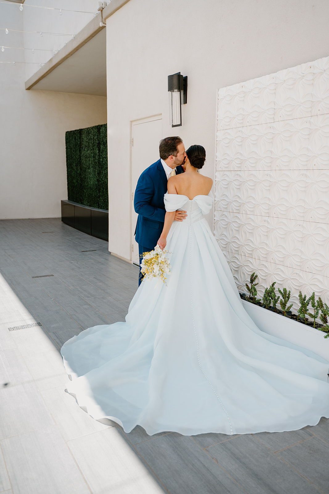 Bride and groom embracing beside a textured white wall, her gown flowing behind her in a romantic spring wedding portrait.