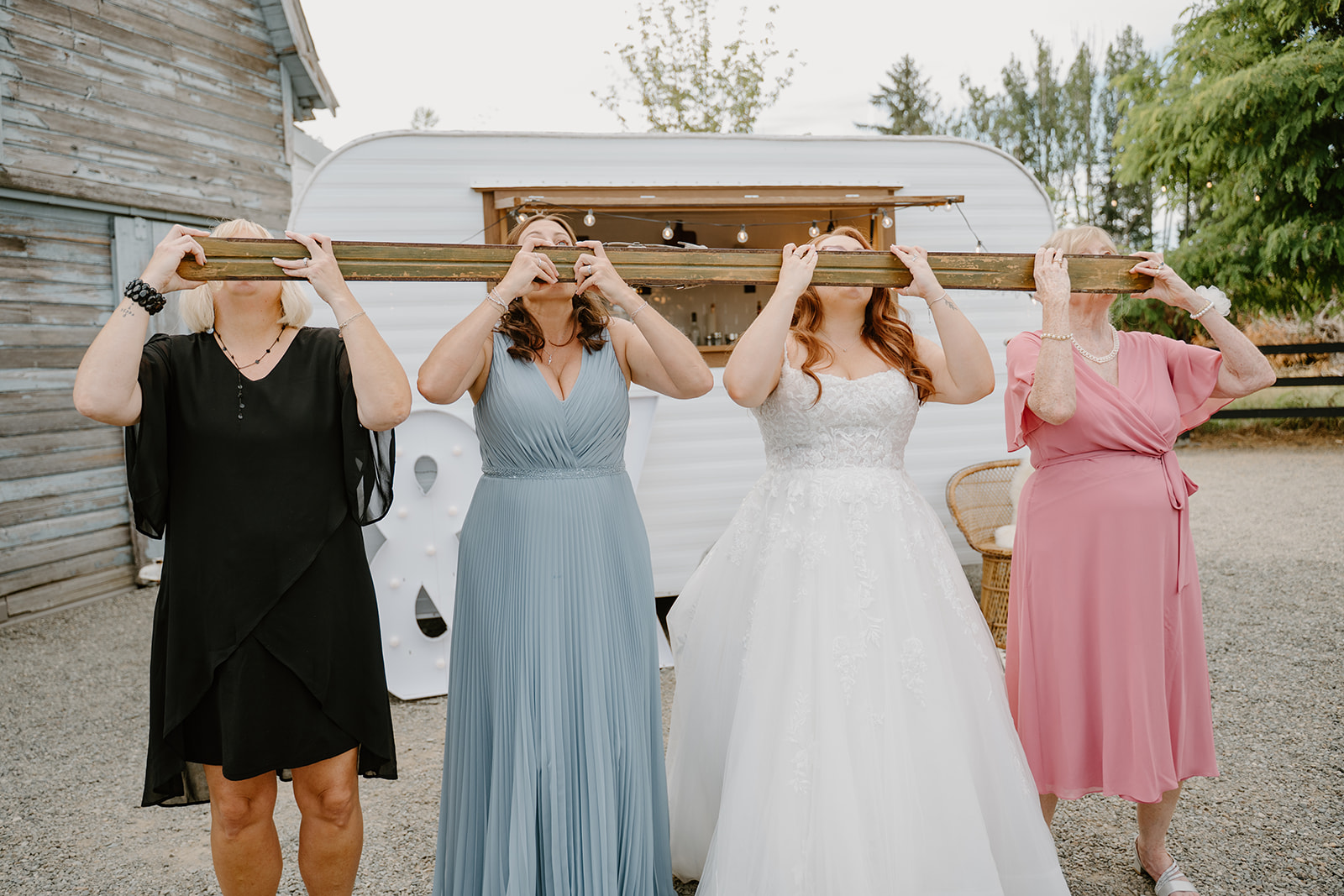 Bride and family members play a fun wedding drinking game using a long wooden board in front of a vintage mobile bar trailer.